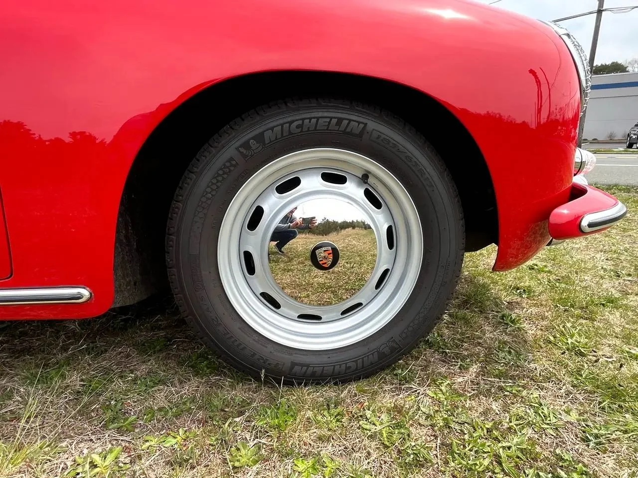 Close-up of a red vintage Porsche car's front wheel with a shiny hubcap reflecting the person taking the photo, parked on grassy ground.
