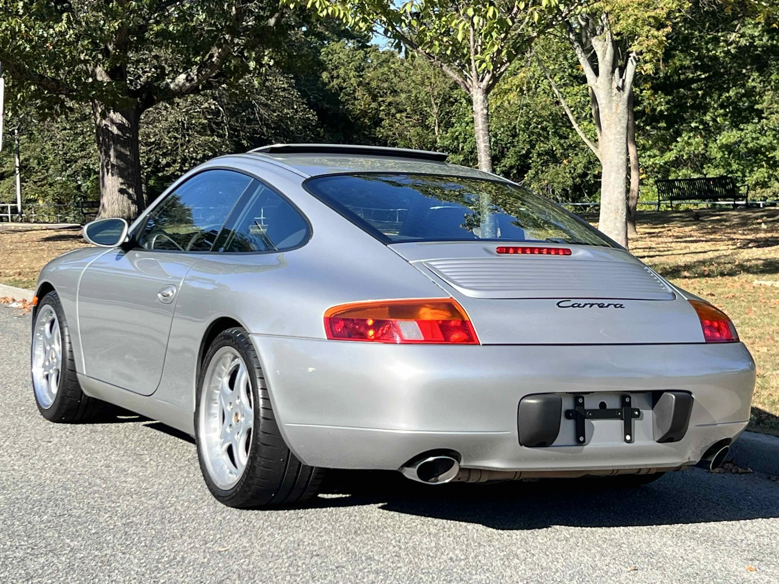 A silver Porsche Carrera sports car parked on a paved surface in a park with trees and benches in the background.