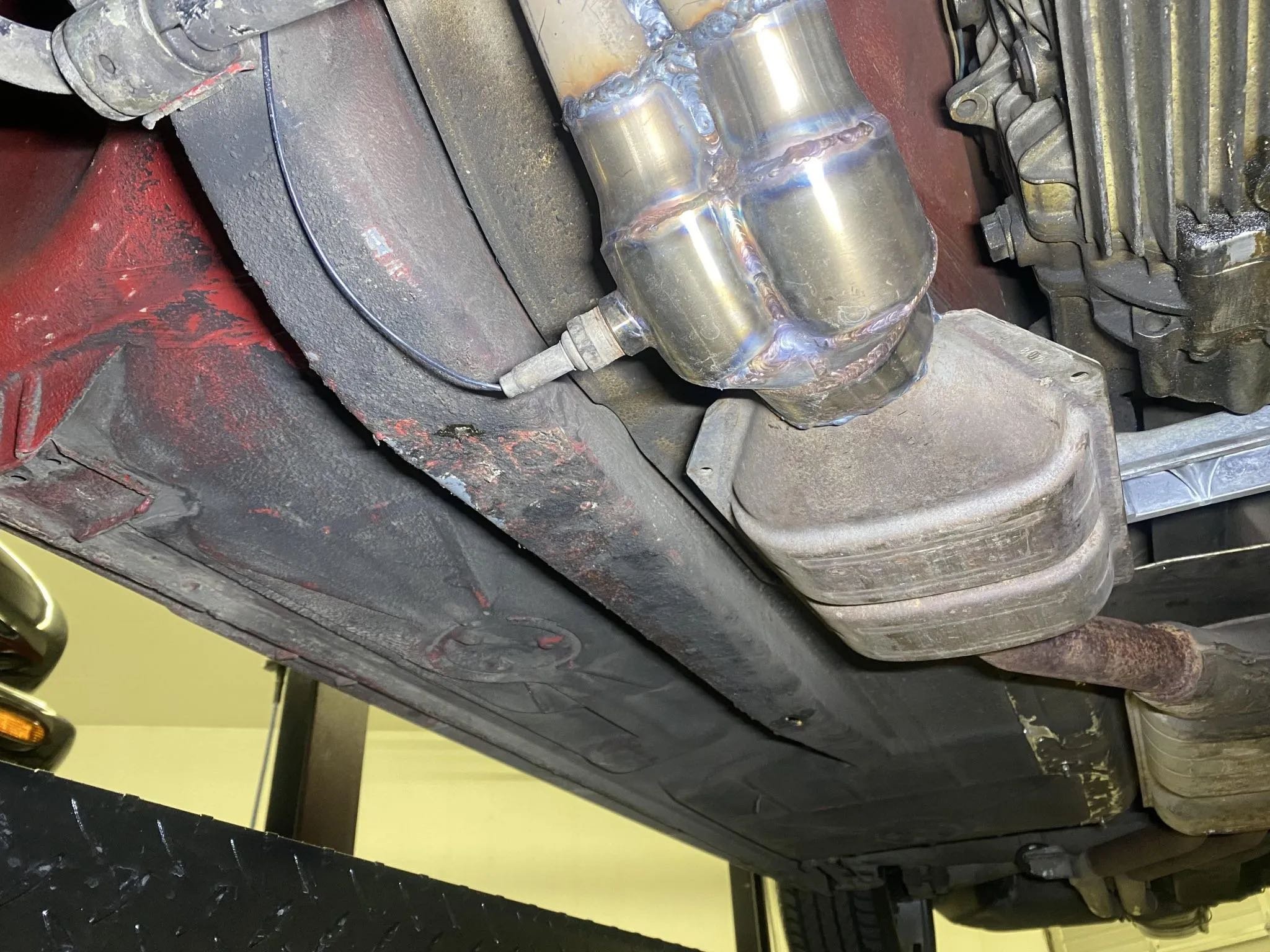 Underneath view of a car's engine and exhaust system showing a metallic oil pan, exhaust pipe, transmission, and rusty parts.