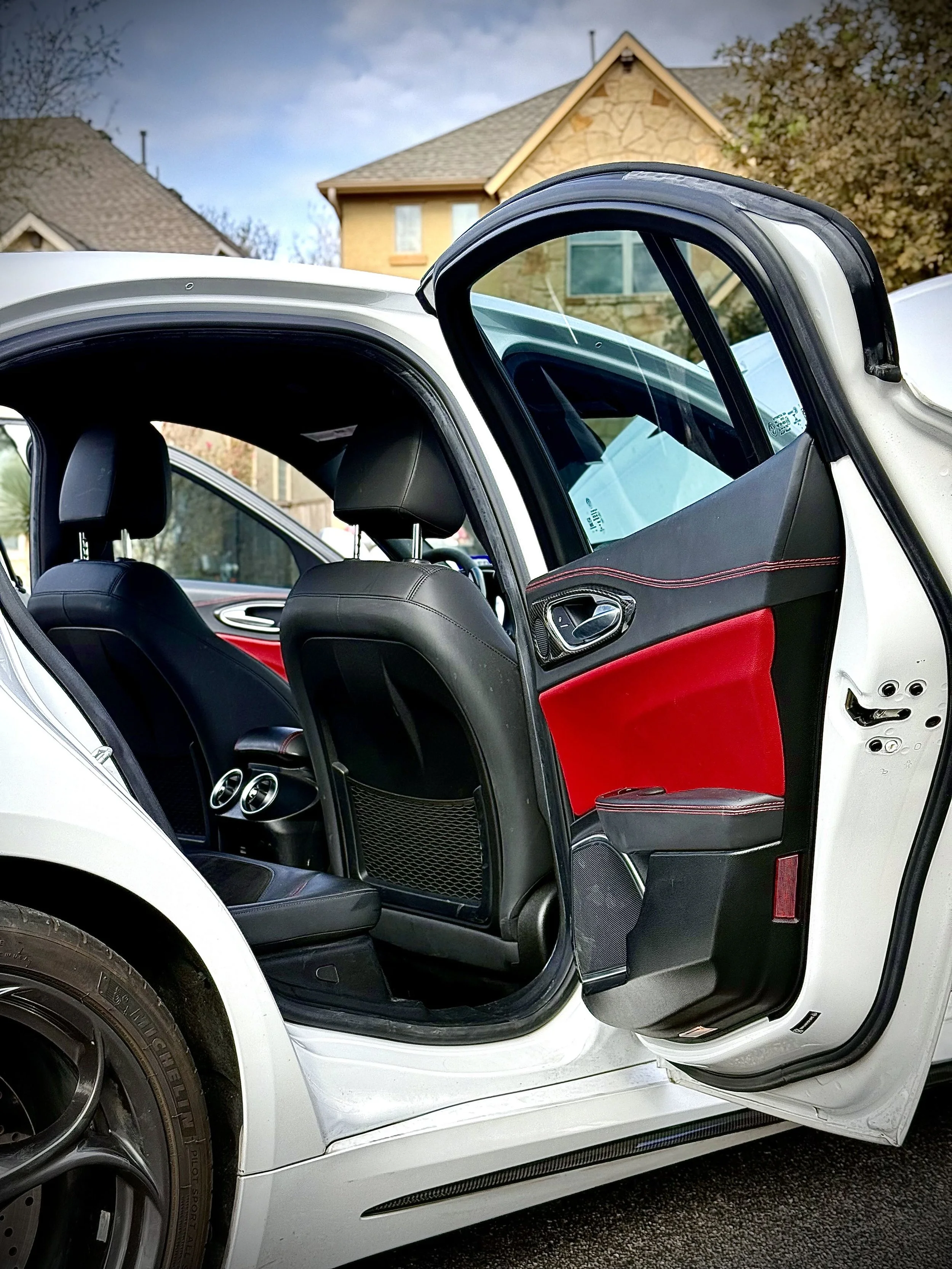 The back door of a white sports car is open, revealing the interior with black and red seats and air vents, with a house visible in the background.