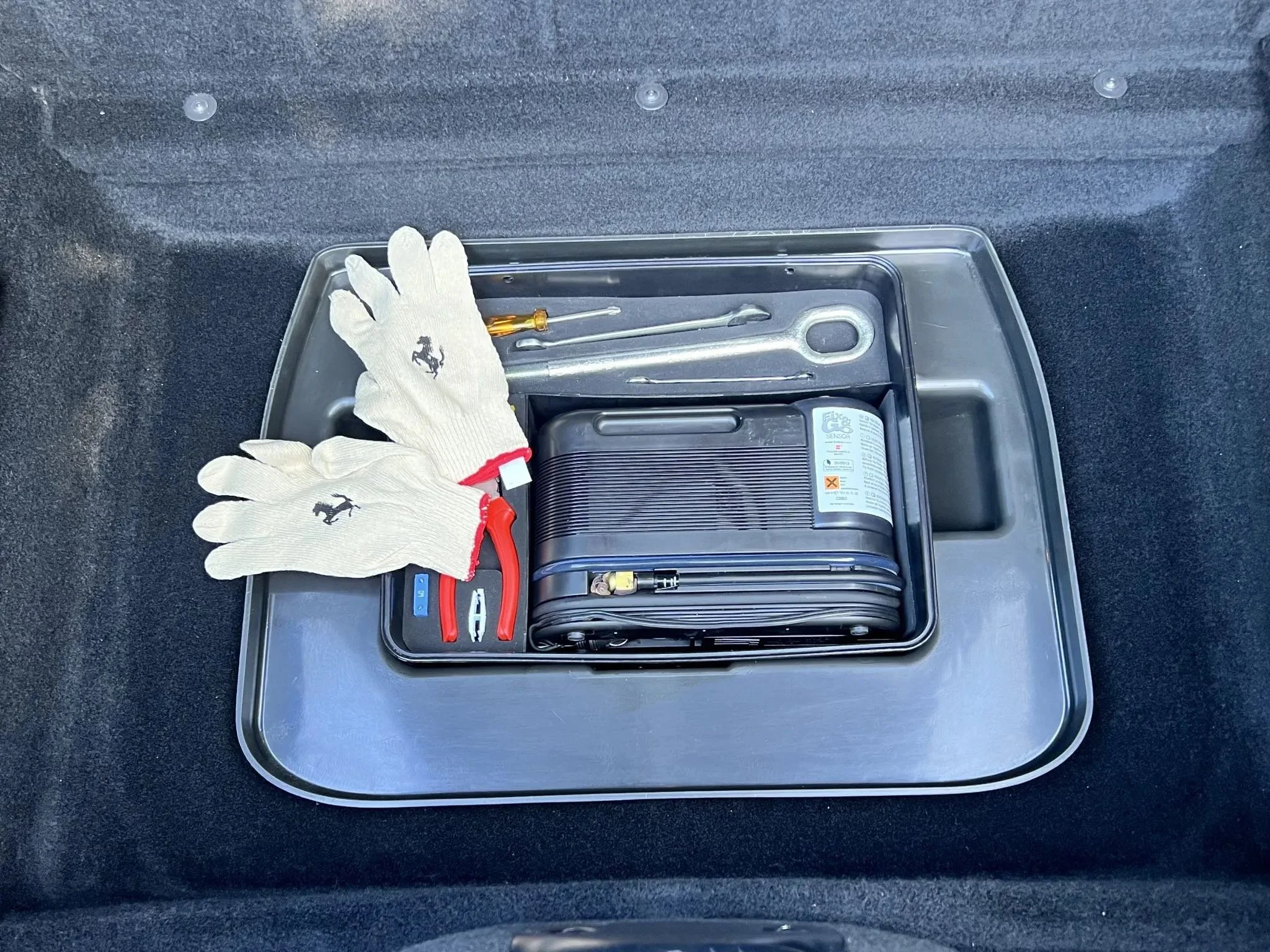 Car trunk with a metal tray containing a folded portable air compressor, work gloves, jumper cables, a screwdriver, and a tire iron.
