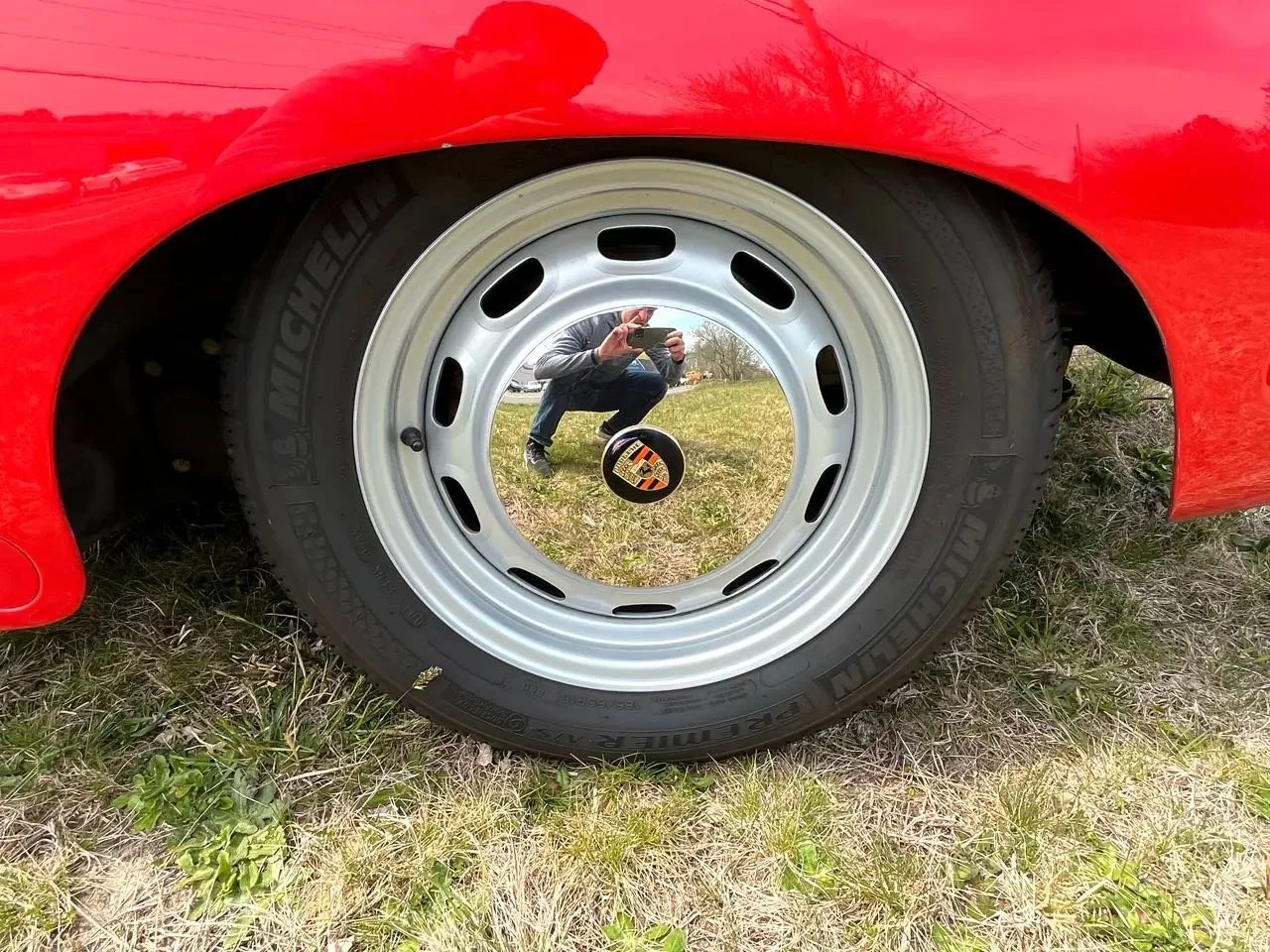A red car with a reflective hubcap, which displays a reflection of a person taking a photo with a phone, in an outdoor grassy area.