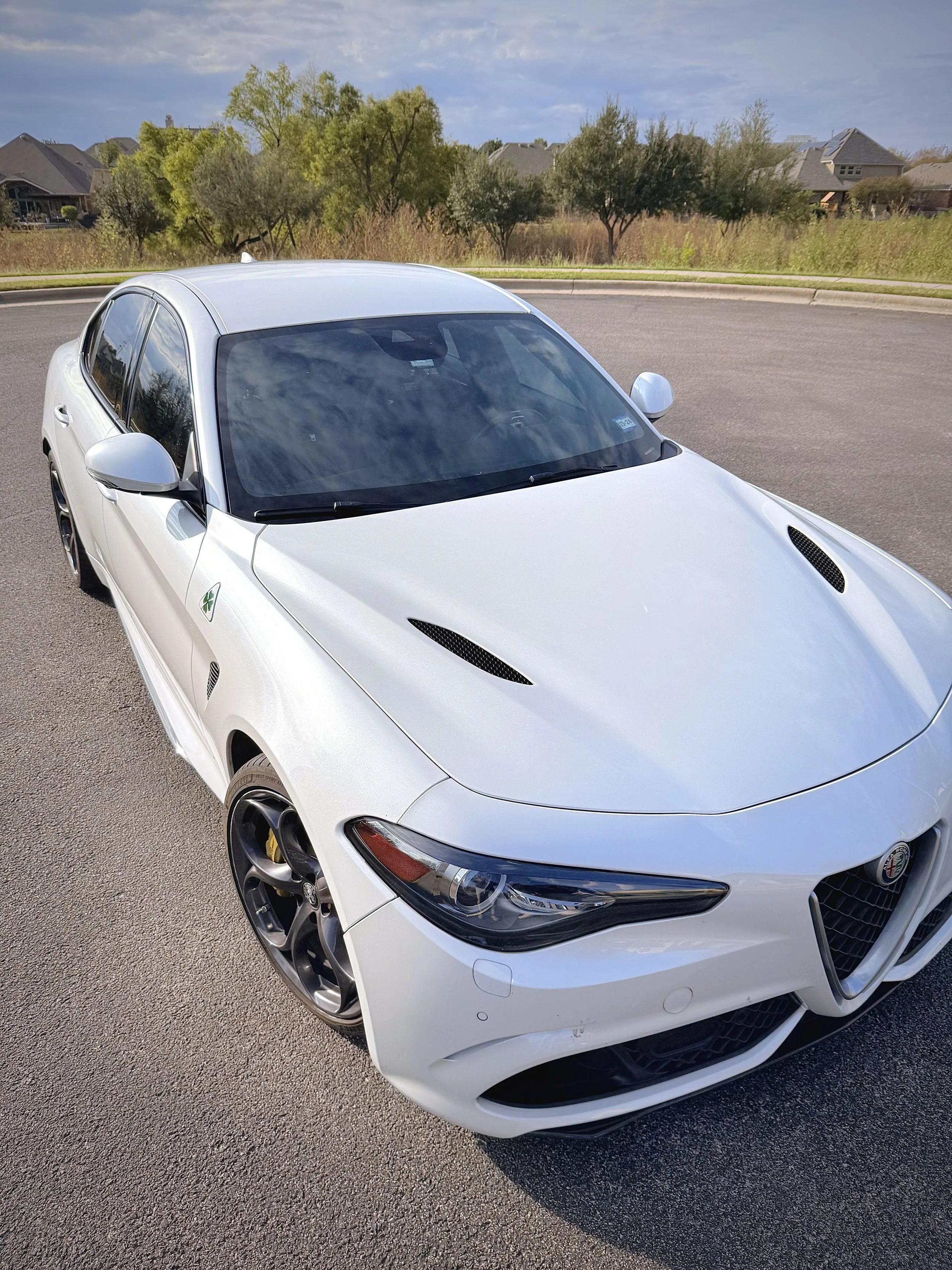A white sports car parked on the side of the road, with trees and suburban houses in the background.