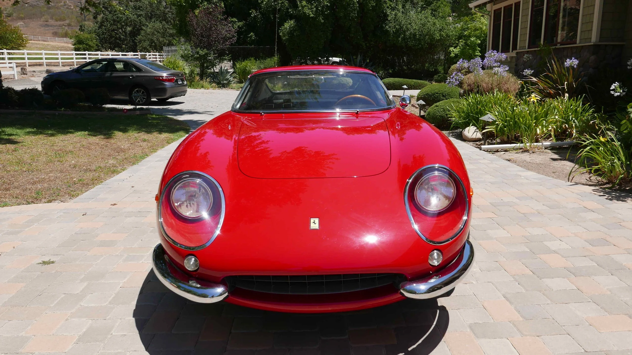 Front view of a red vintage Ferrari sports car parked on a brick driveway, with a black modern sedan in the background and a garden with plants and flowers to the side.