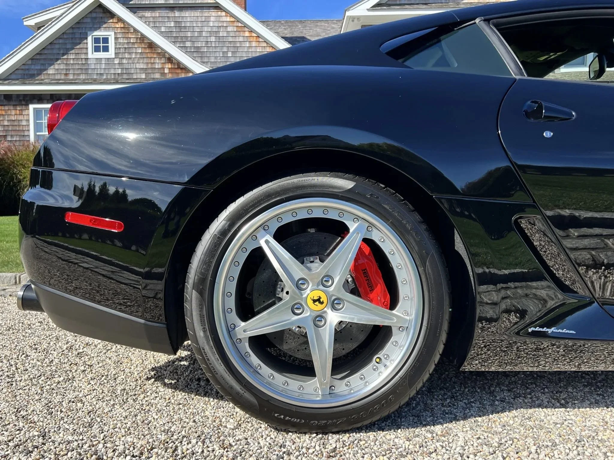 Close-up of the rear wheel of a black Ferrari, showing a silver, five-spoke rim with the yellow Ferrari logo, red brake calipers, and a drilled brake disc.