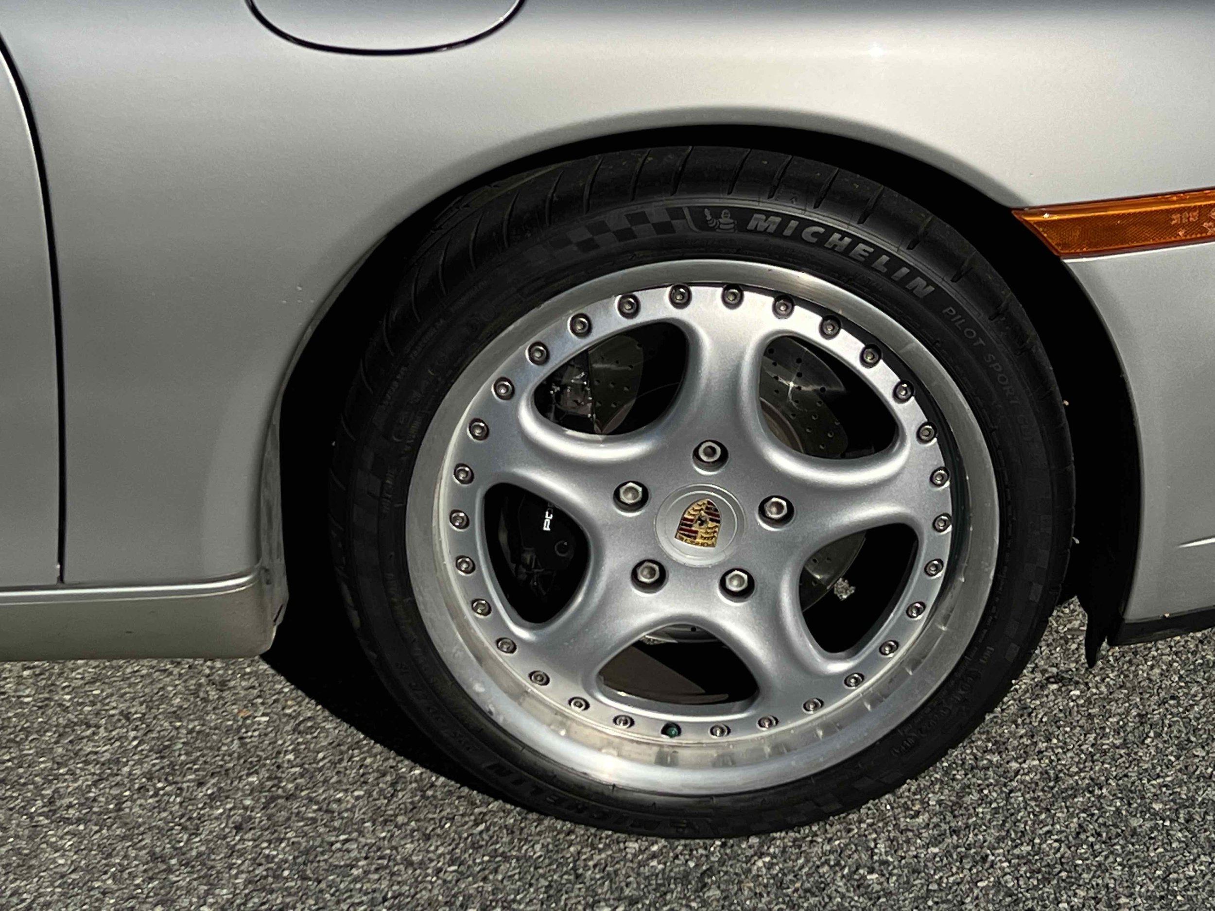 A silver car with a close-up of a wheel featuring a Porsche emblem, black Michelin Pilot Sport tire, and polished multi-piece rim with rivets.