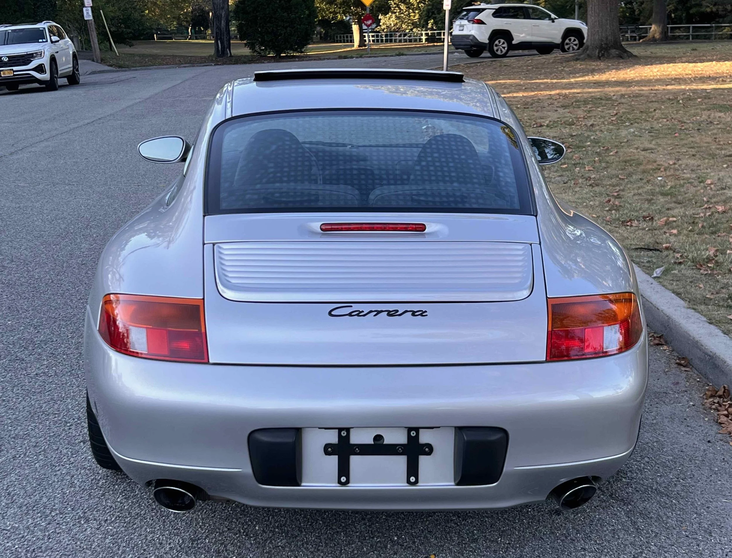 Rear view of a silver Porsche Carrera parked on the side of a road, with other vehicles in the background, and a grassy area with trees.