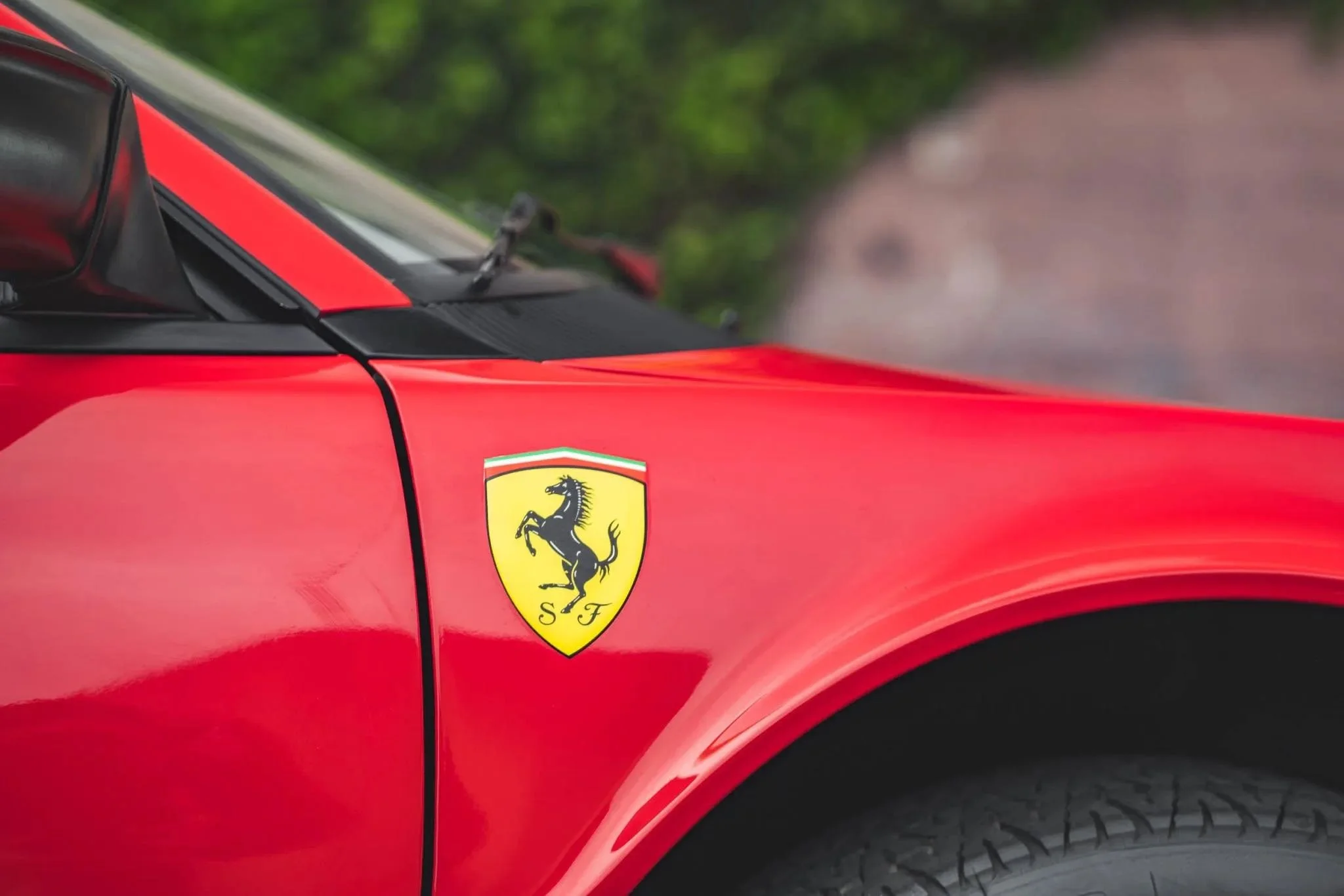 Close-up of a red Ferrari car with the Ferrari logo on the side, showing part of the front wheel and fender, against a blurred outdoor background.