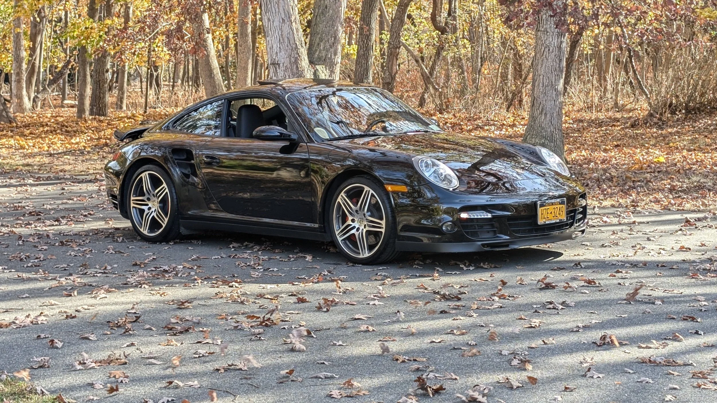 A black Porsche sports car parked on a leaf-covered road in a wooded area during fall.