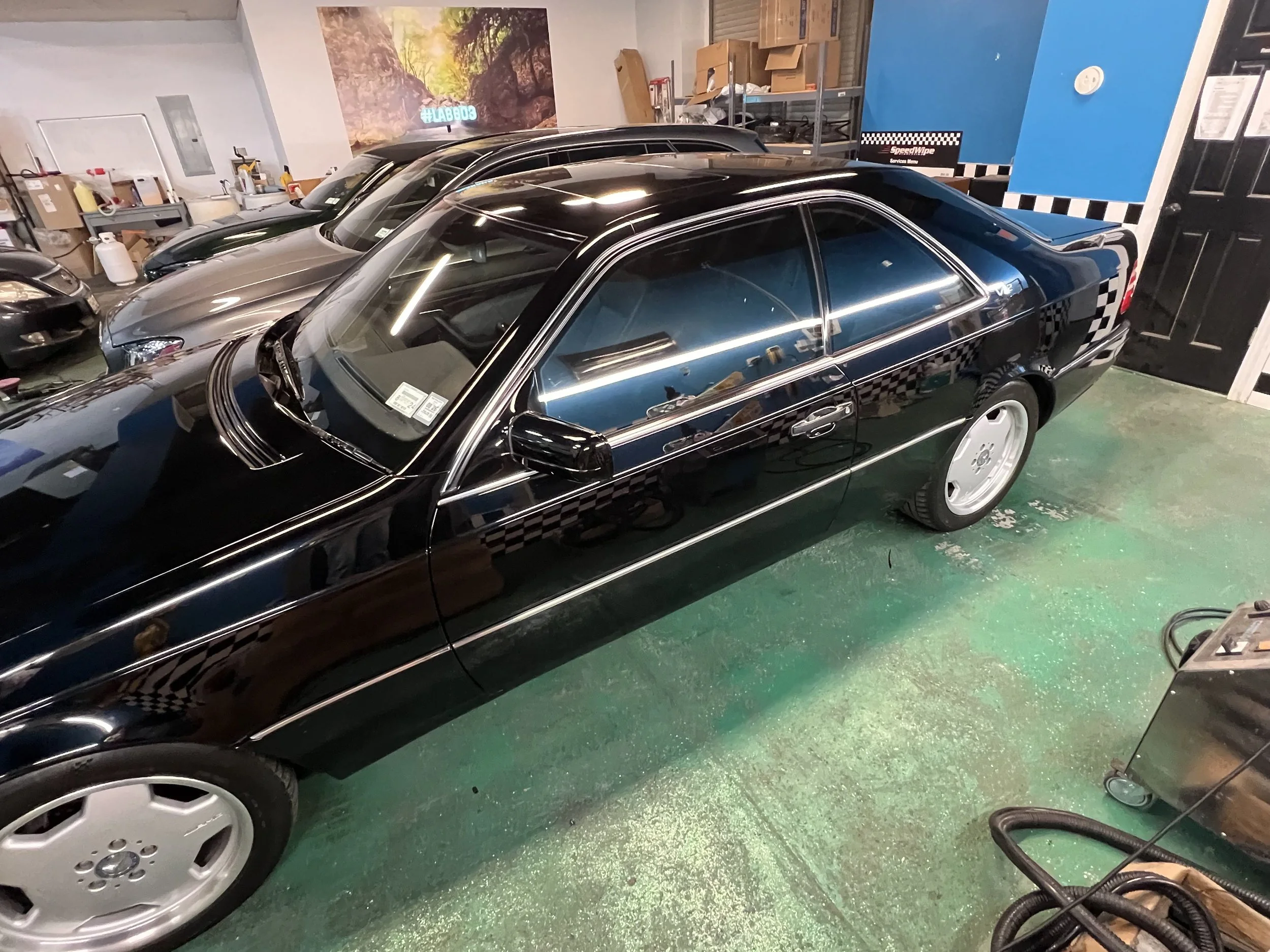 Black vintage Mercedes-Benz car parked inside a garage alongside other vehicles, with a checkered pattern on the side and rear window.