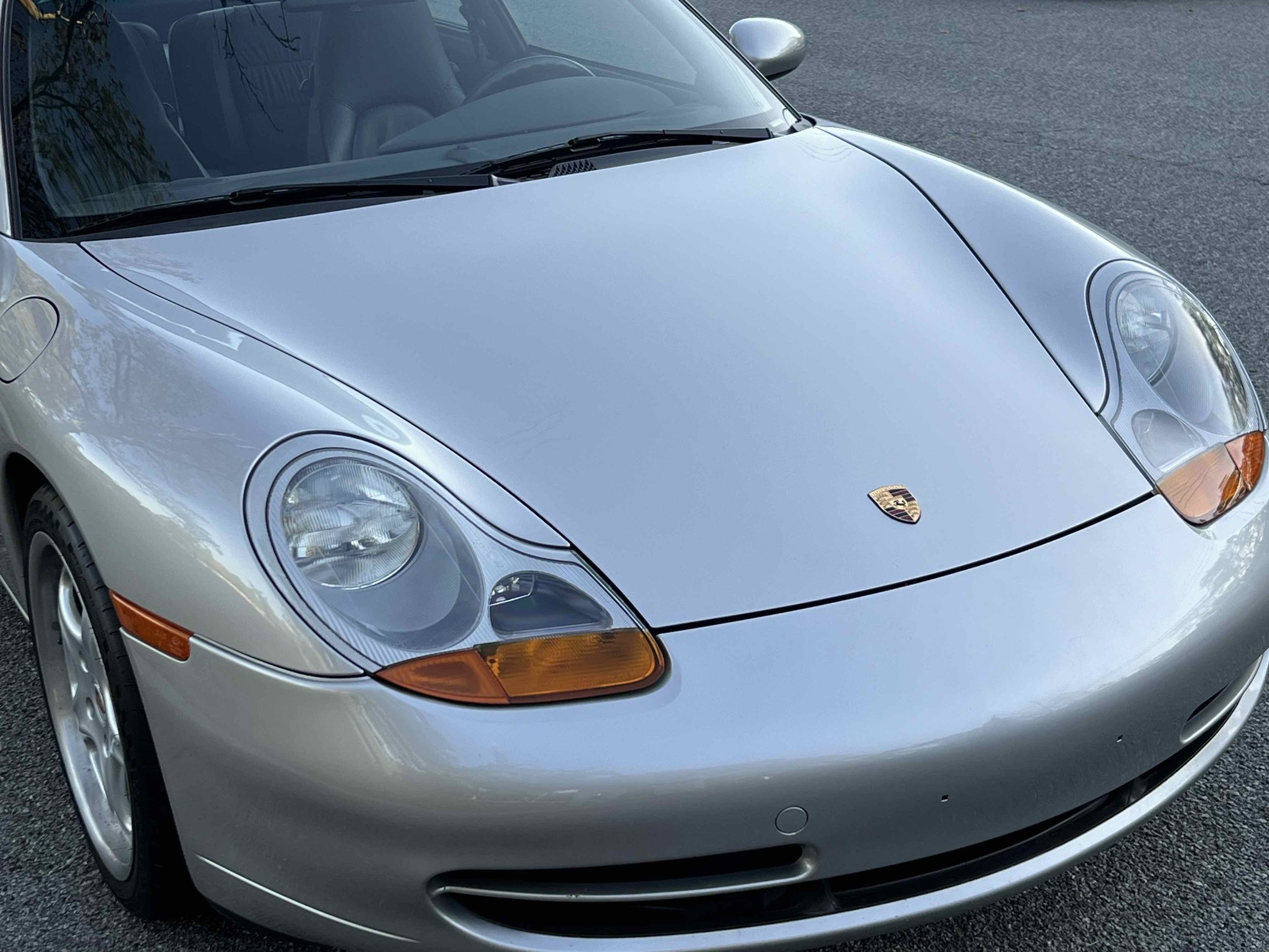 Close-up of front of a silver Porsche sports car parked on a paved surface, showing the headlights, hood, and the Porsche emblem.