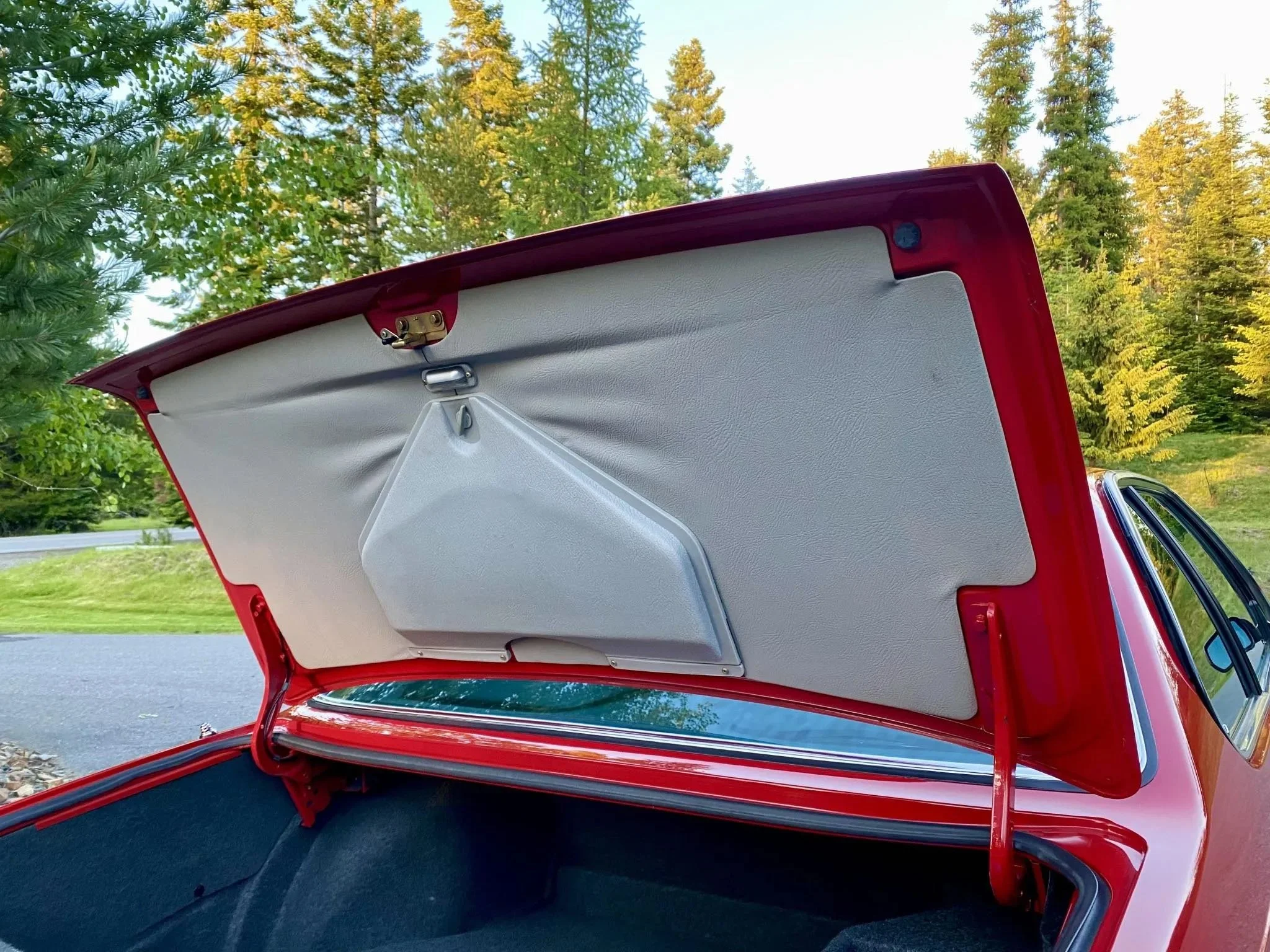 Open trunk of red vintage car outdoors with trees and sky in background.