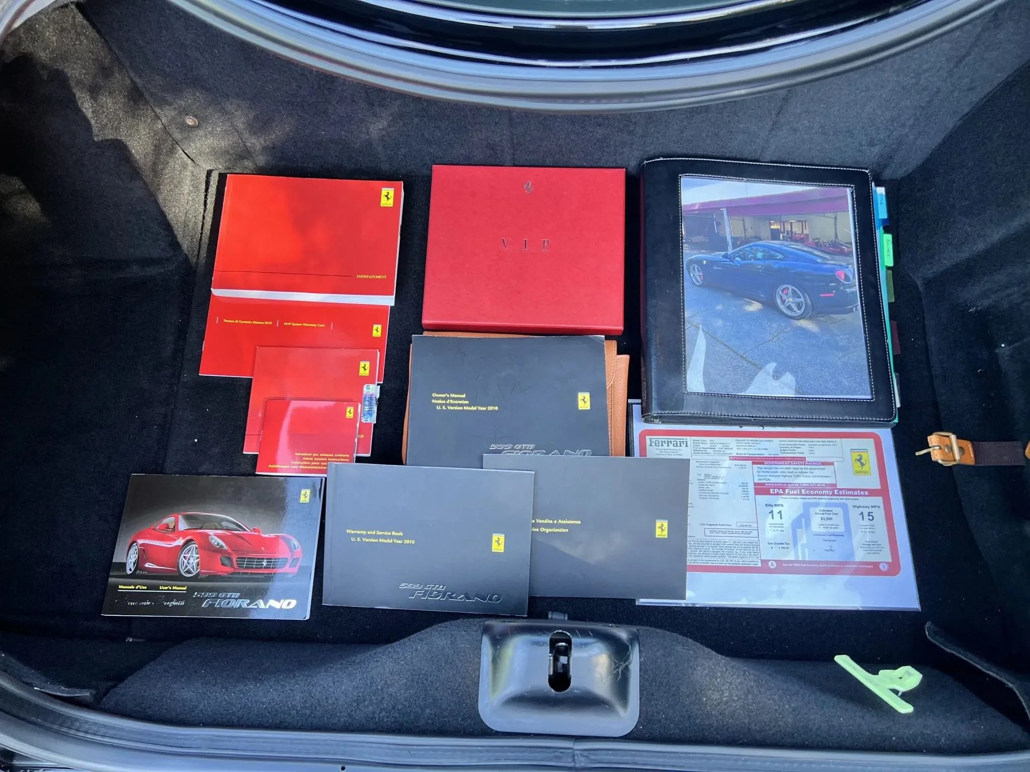 Collection of Ferrari manuals, brochures, and documents laid out in a car trunk, including a red Ferrari brochure featuring a red sports car, a black owner's manual, a red cover, a black service book, various booklets, and a document with fuel econom