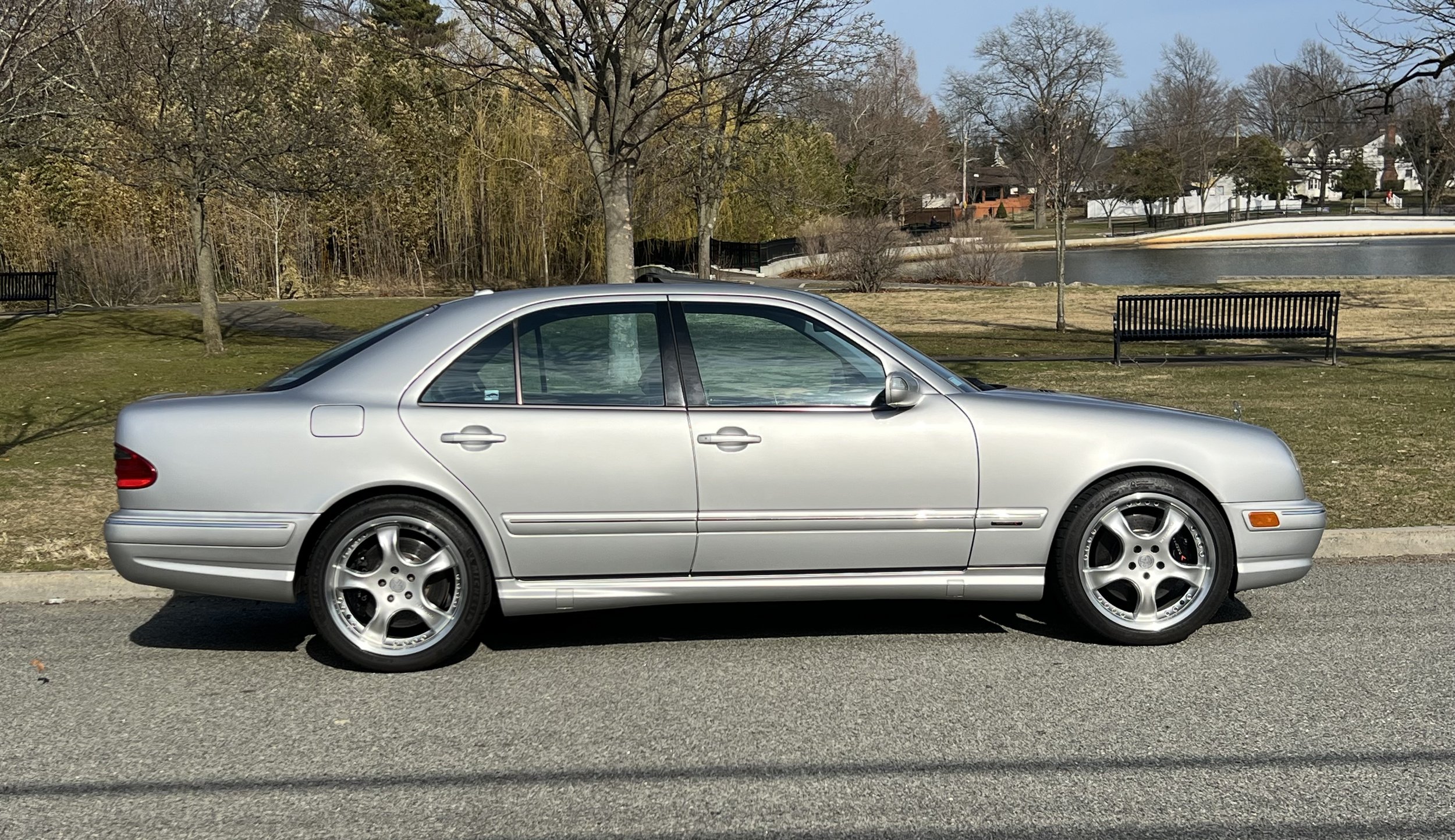 A silver sedan car parked on the side of a street with trees, benches, and a body of water in the background.