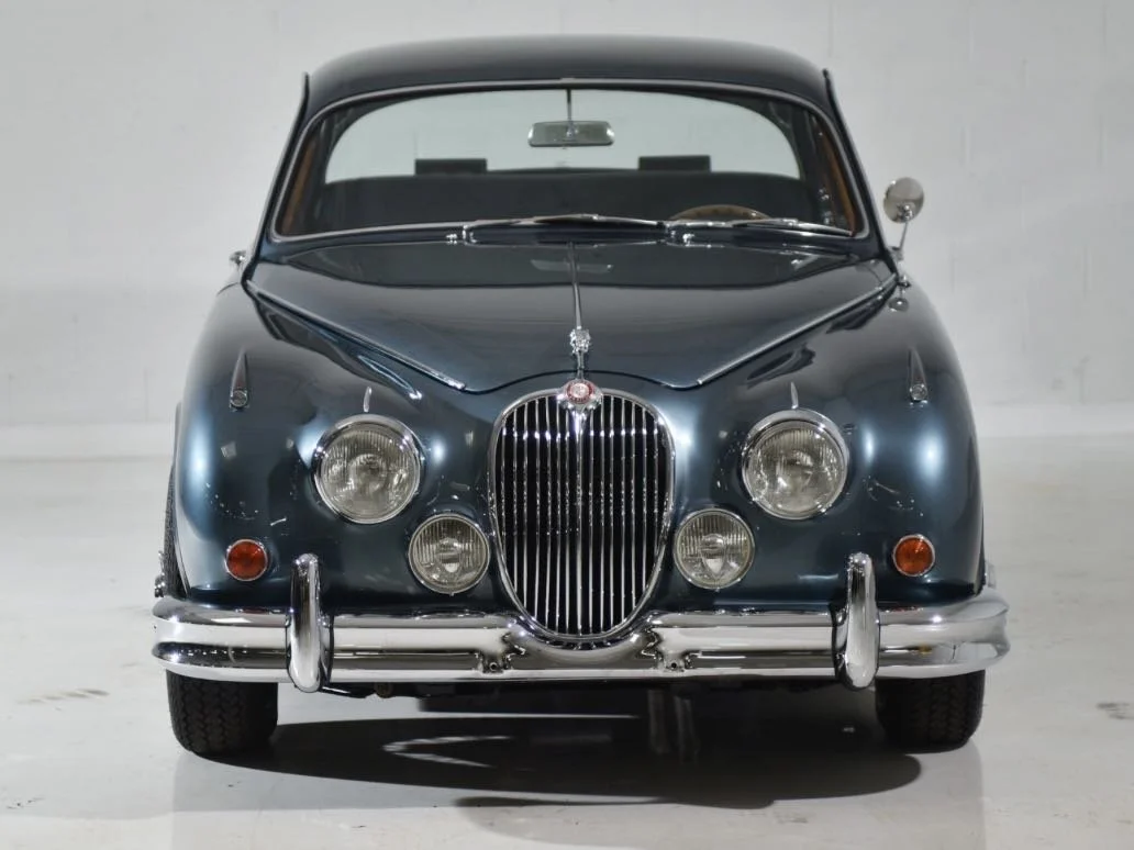 Front view of a vintage dark blue classic car with a distinctive grille, four headlights, and chrome bumper, parked against a plain white background.