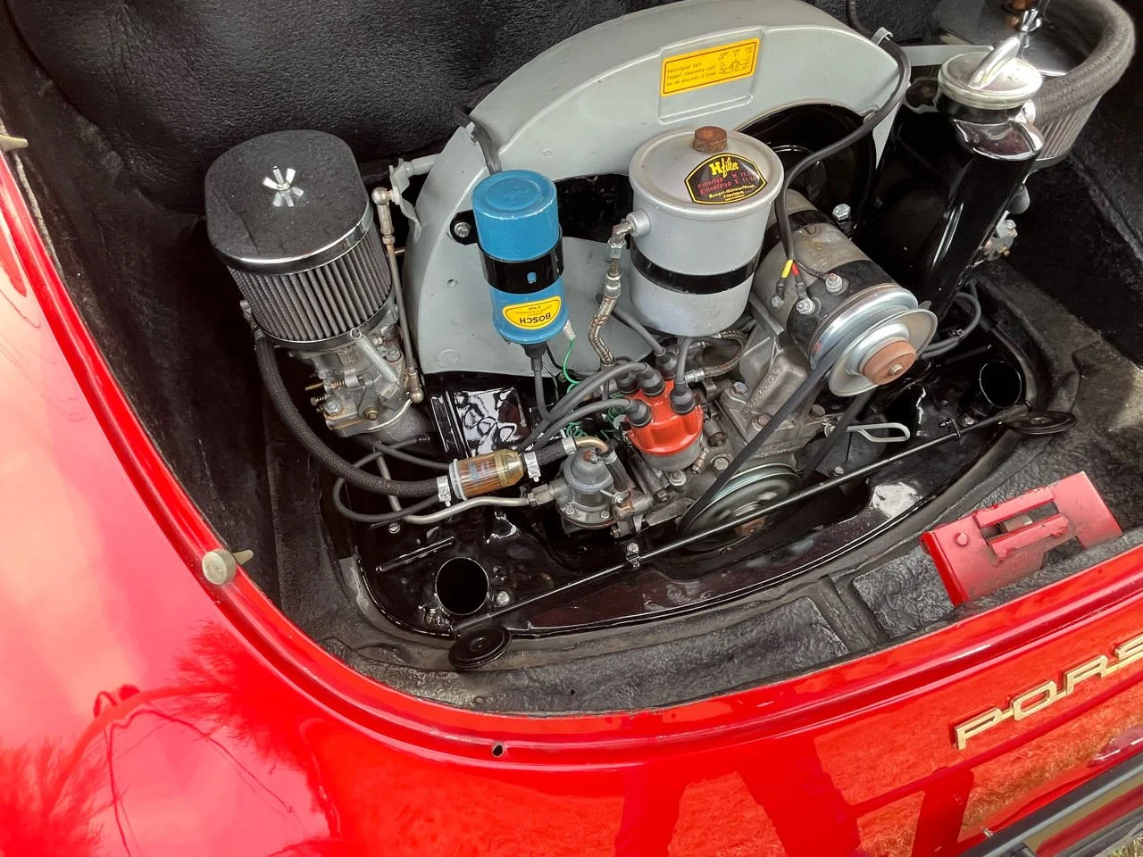 The image shows the engine compartment of a classic red Porsche sports car, featuring a detailed view of the engine with various components, wires, and a Bosch blue oil filter.