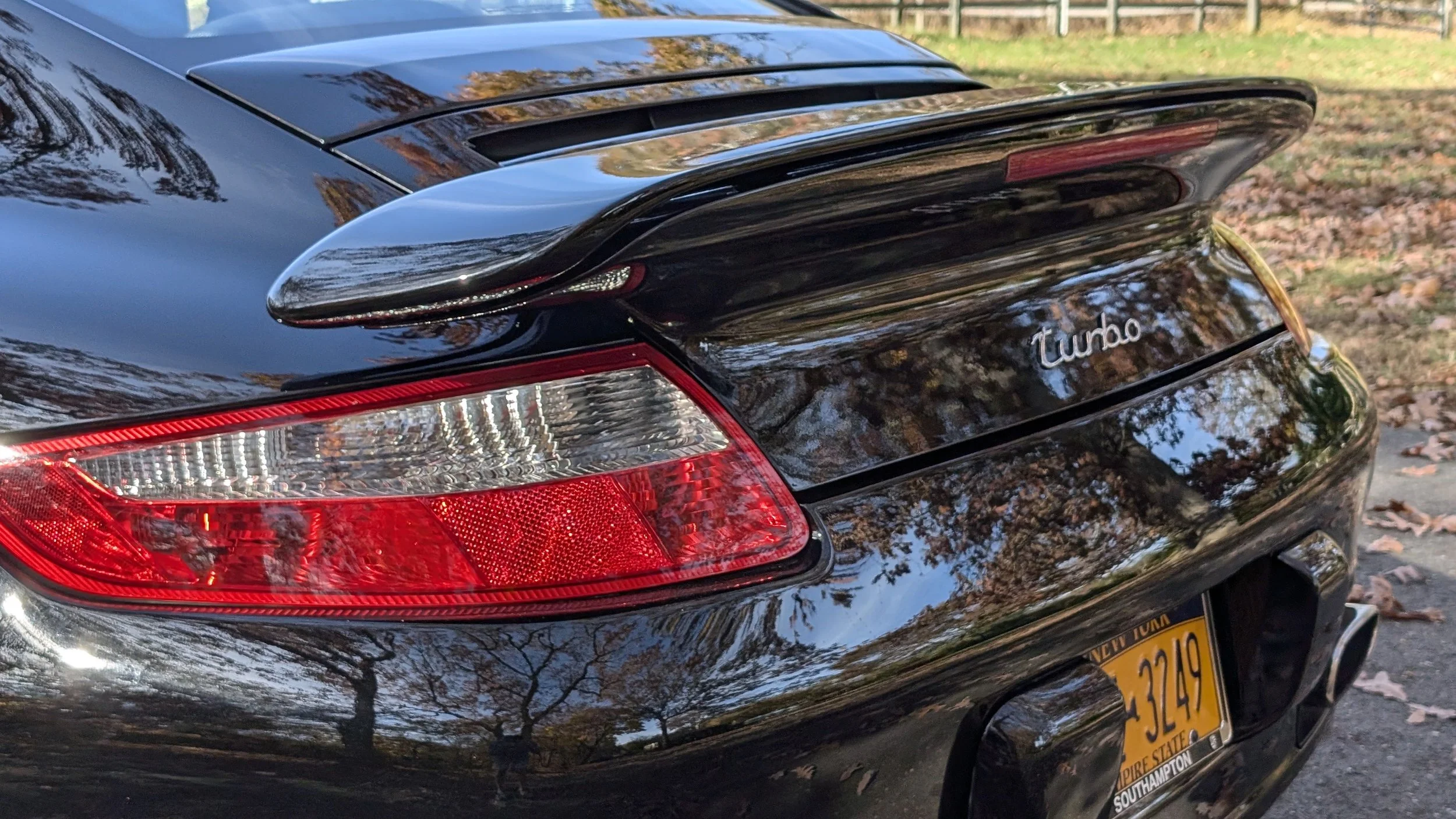 Rear view of a black Porsche 911 Turbo with a spoiler, showing taillights and a New York license plate, parked on a tree-lined street with fallen leaves.