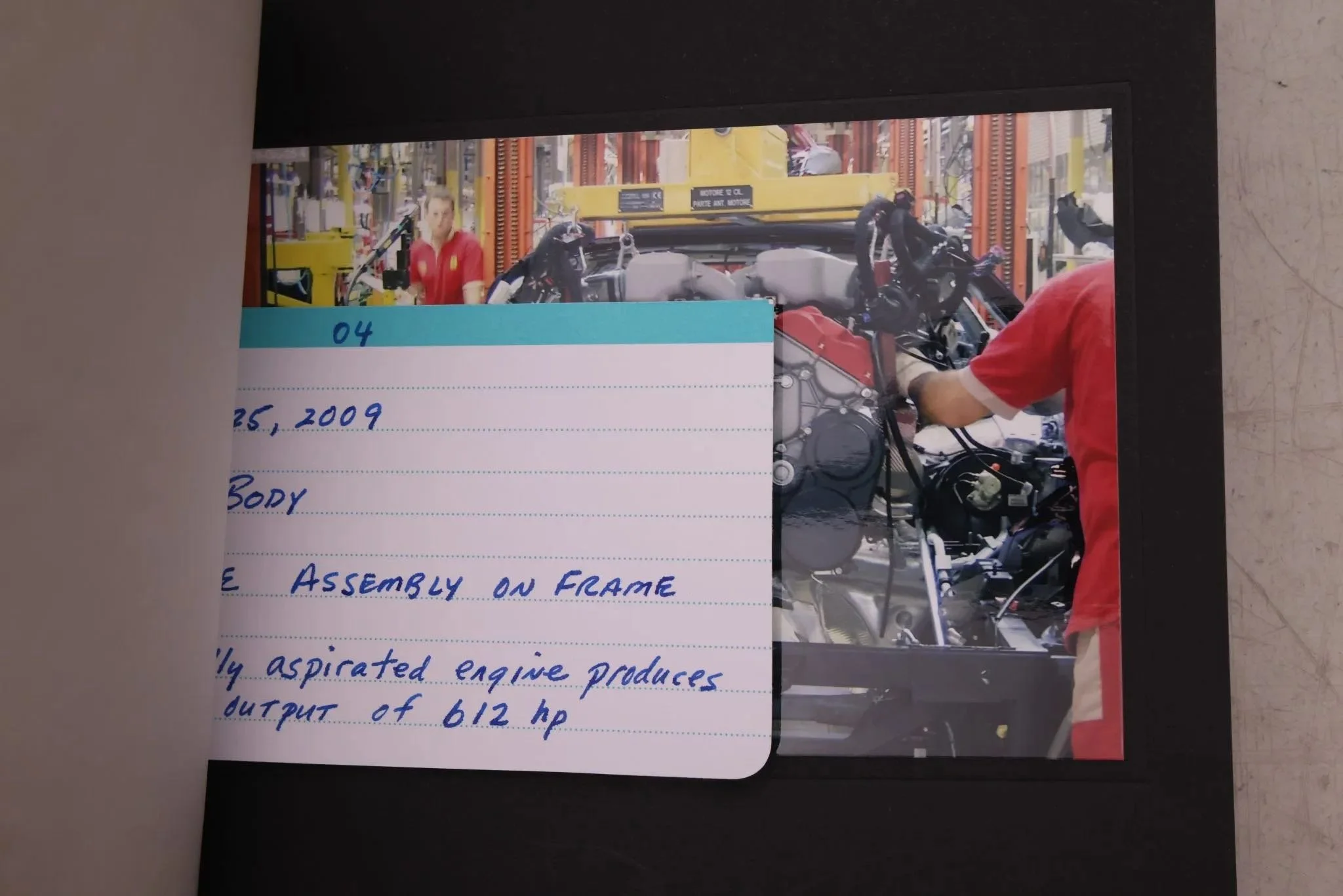 Photo of a mechanic working on a motorcycle engine in a workshop, with a person in a red shirt visible on the right side of the image, and tools and equipment surrounding the workspace.