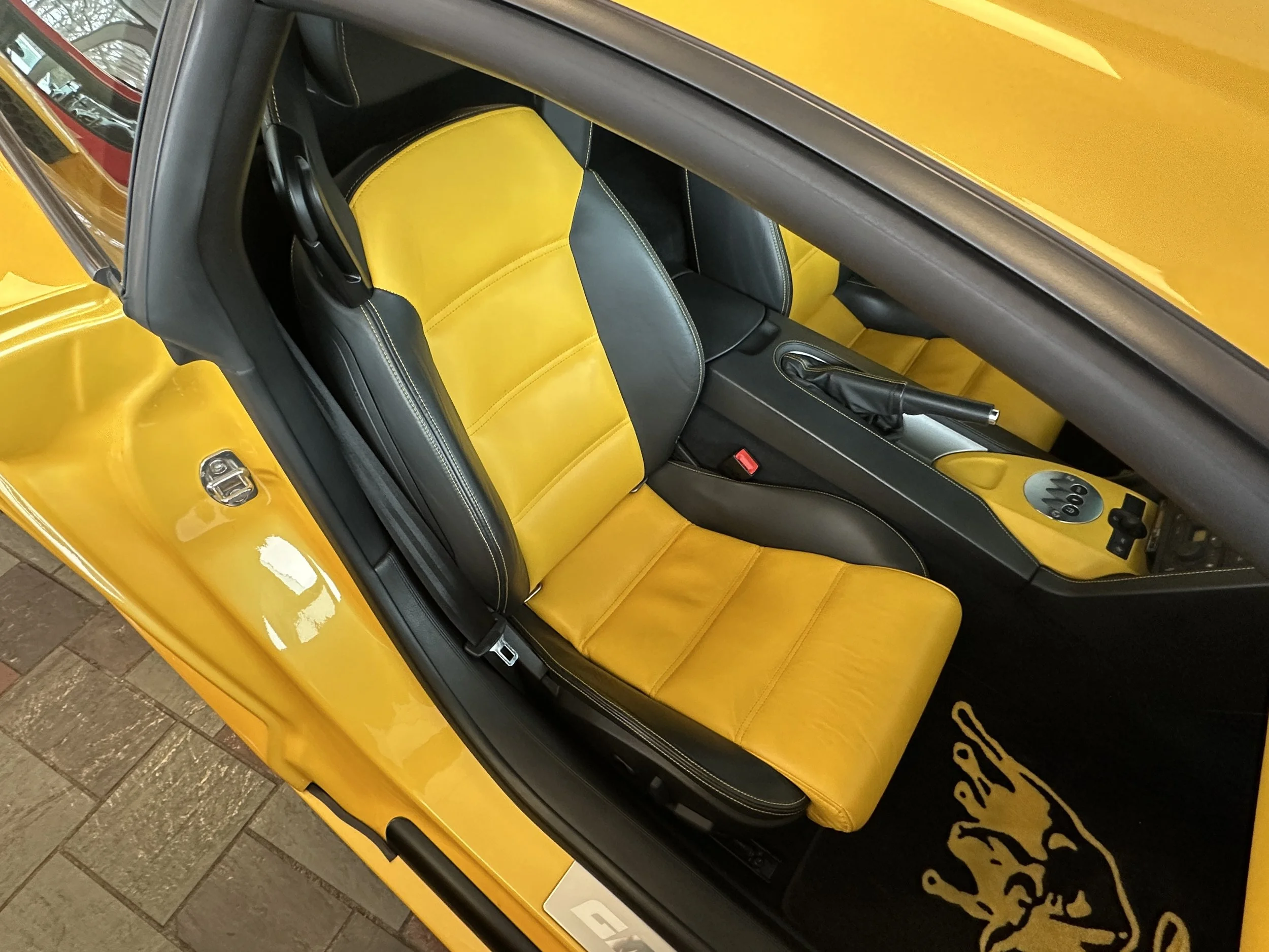 Inside a yellow sports car, showing black and yellow leather seats, the gear shift, and a black floor mat with a yellow horse logo.