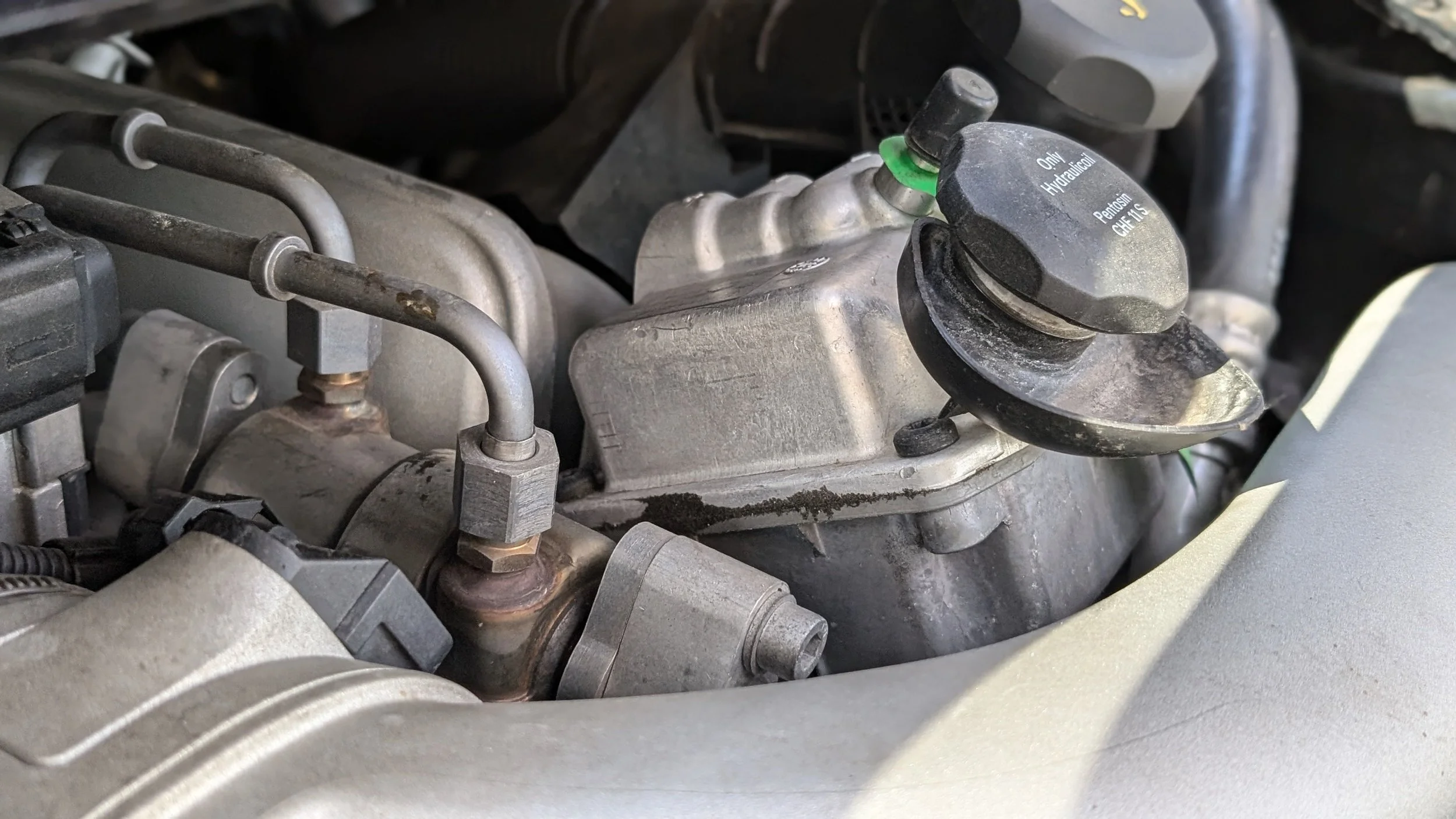 Close-up of car engine components, including fuel injectors, metal pipes, and valve cover, with some dirt and grime.