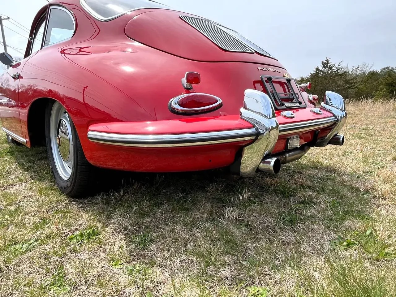 Rear view of a vintage red Porsche 356 with chrome bumpers, taillights, and exhaust pipe, parked on grass.