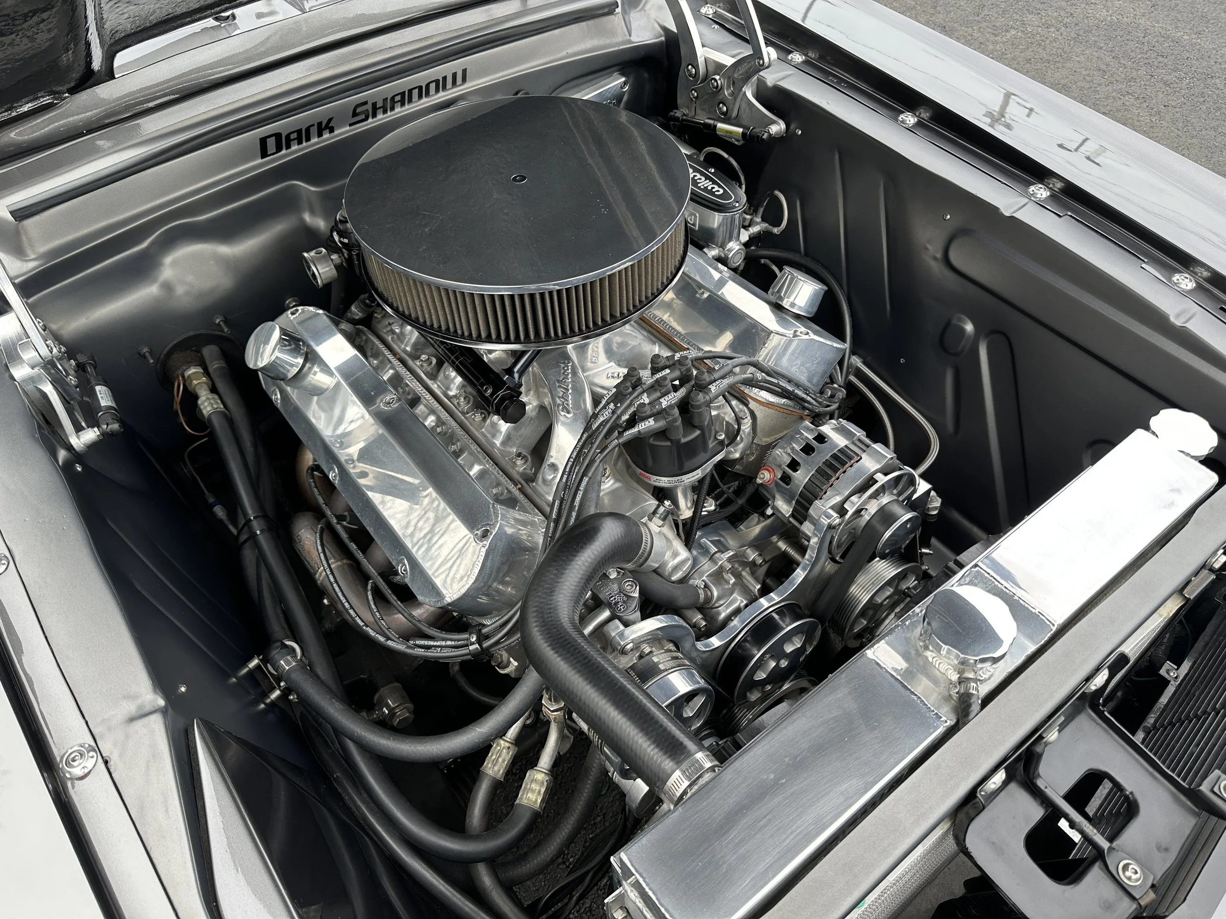 Close-up of a car engine in a classic vehicle, featuring a large round air filter, shiny metallic components, black hoses, and a detailed engine bay with the words 'Dark Shadow' written on the inner panel.