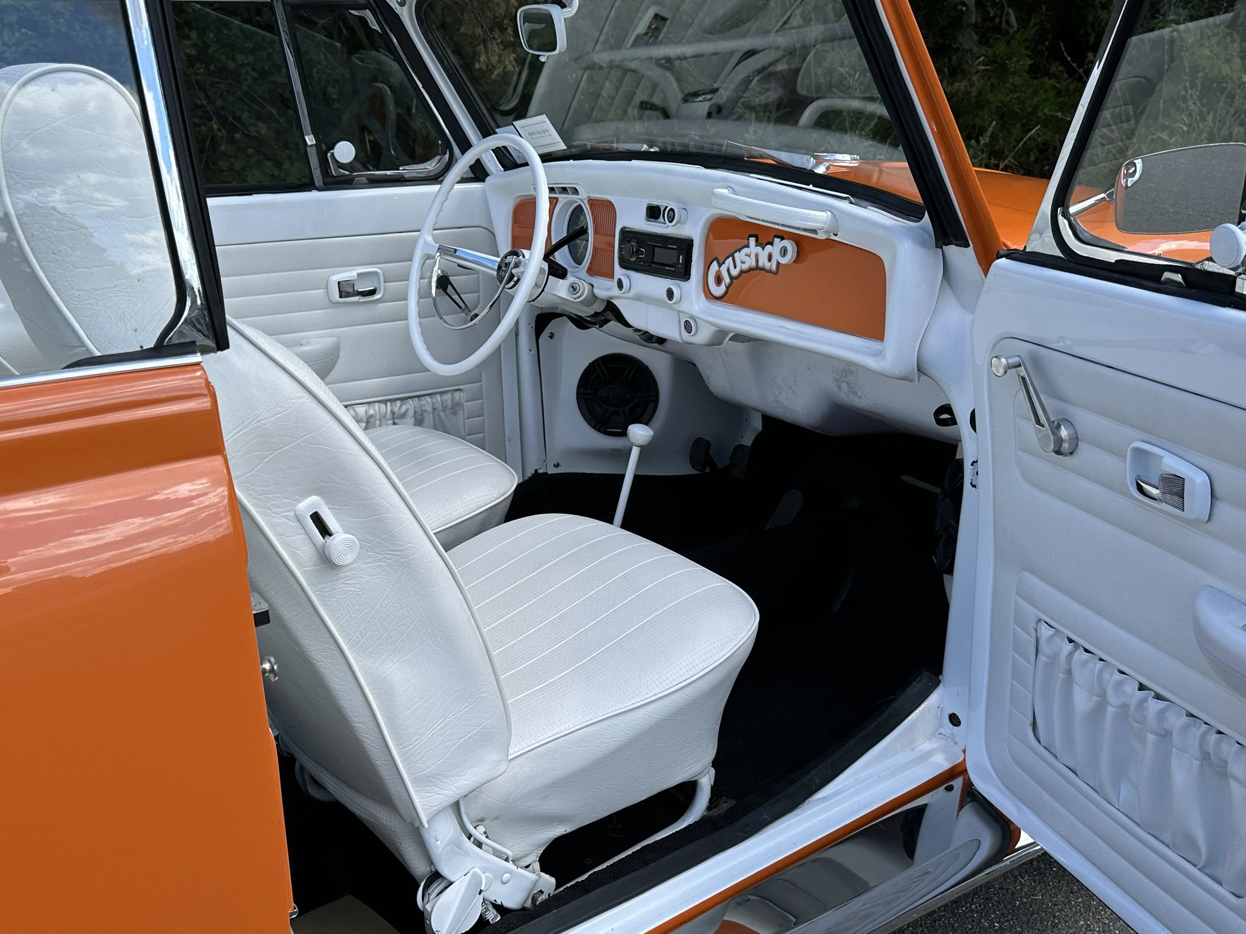 The interior of a vintage Volkswagen Type 2 van with white seats and dashboard, orange accents, and a 'Crushup' sticker on the glove compartment. Orange exterior visible through the open door.