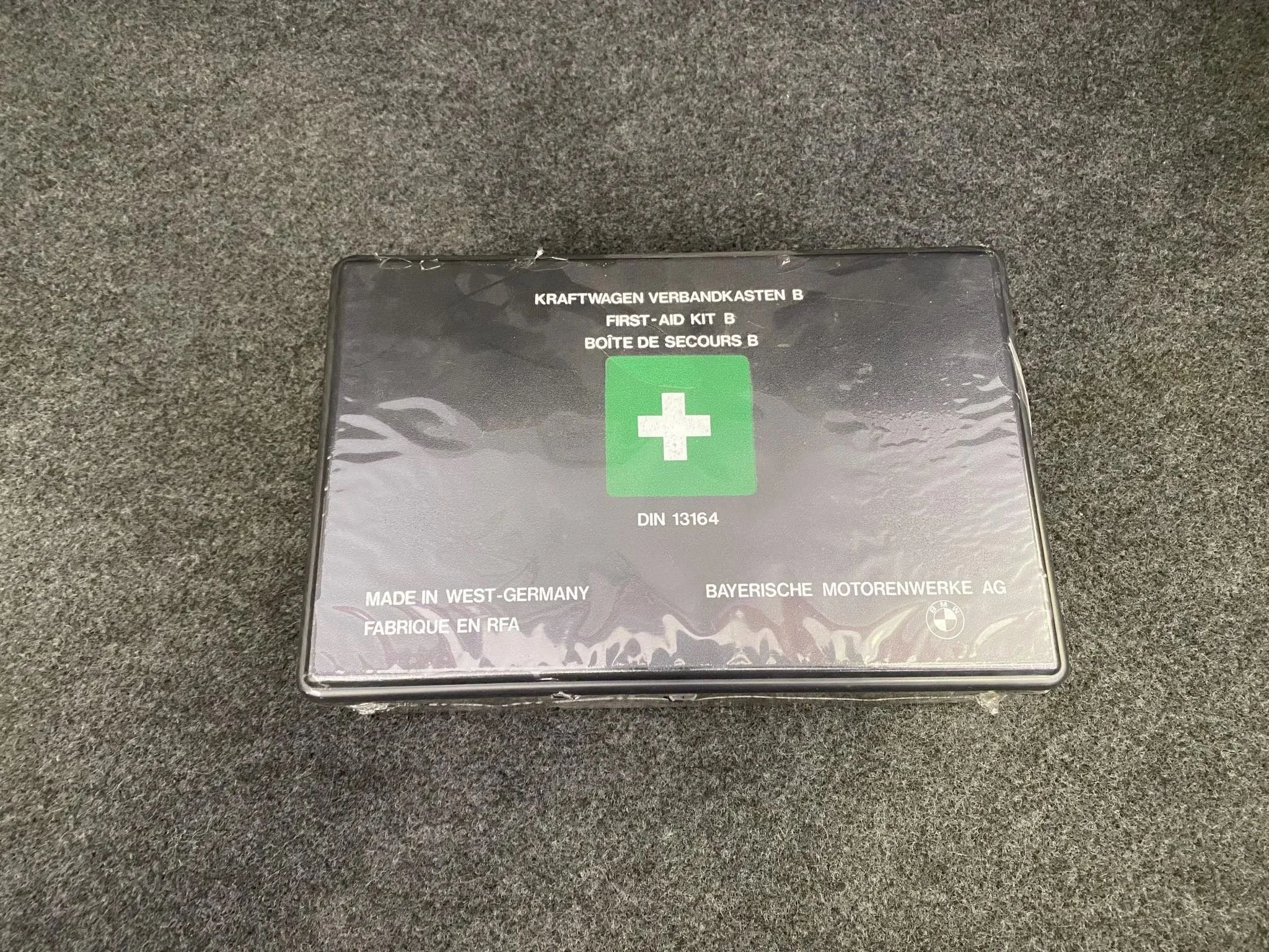 A boxed first aid kit with German and English labels on a dark textured surface.