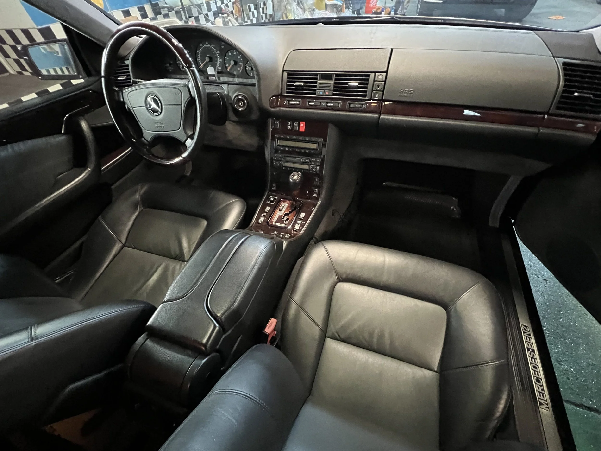 The interior of a black Mercedes-Benz car, featuring a leather driver's seat, steering wheel, and dashboard with various controls and dials. The interior has wood accents and a center console with gear shift and buttons.