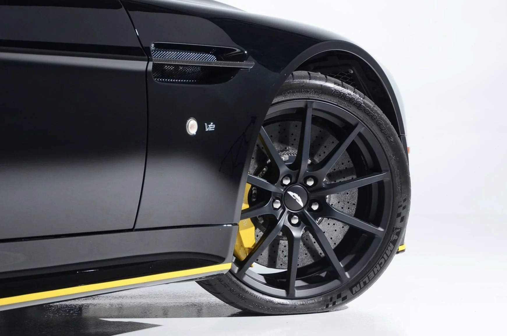 Close-up of a black sports car showing part of the front wheel and tire, with a distinctive yellow brake caliper, a black multi-spoke rim, and a tire with visible branding, against a white background.