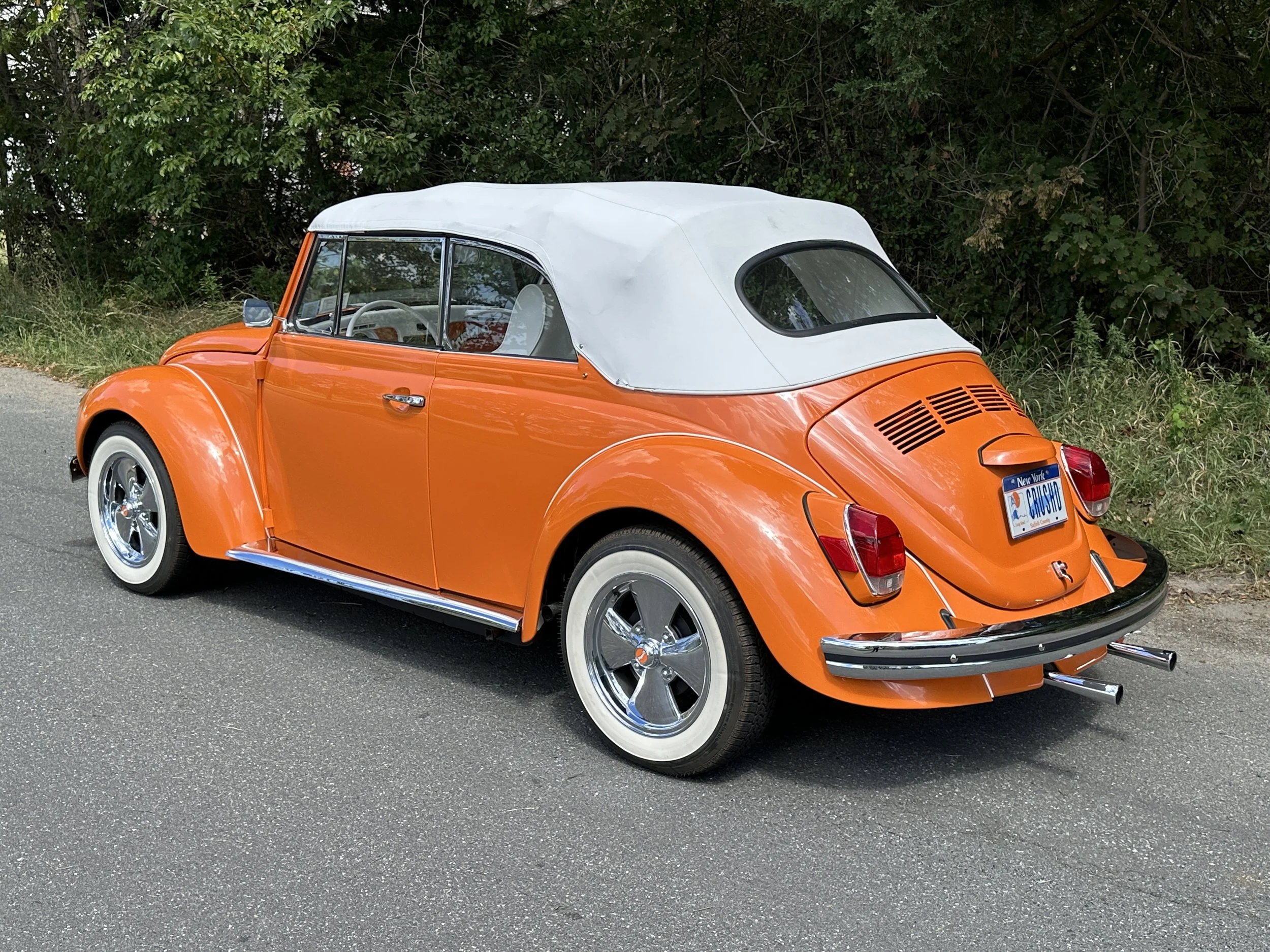 Orange vintage Volkswagen Beetle convertible with a white soft top parked on the side of a road next to a grassy area with trees.