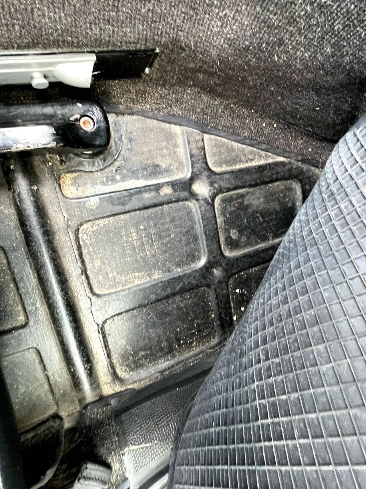 Dirty car floor carpet and rubber floor mat, with a small section of a car seat in a silver and black pattern visible.