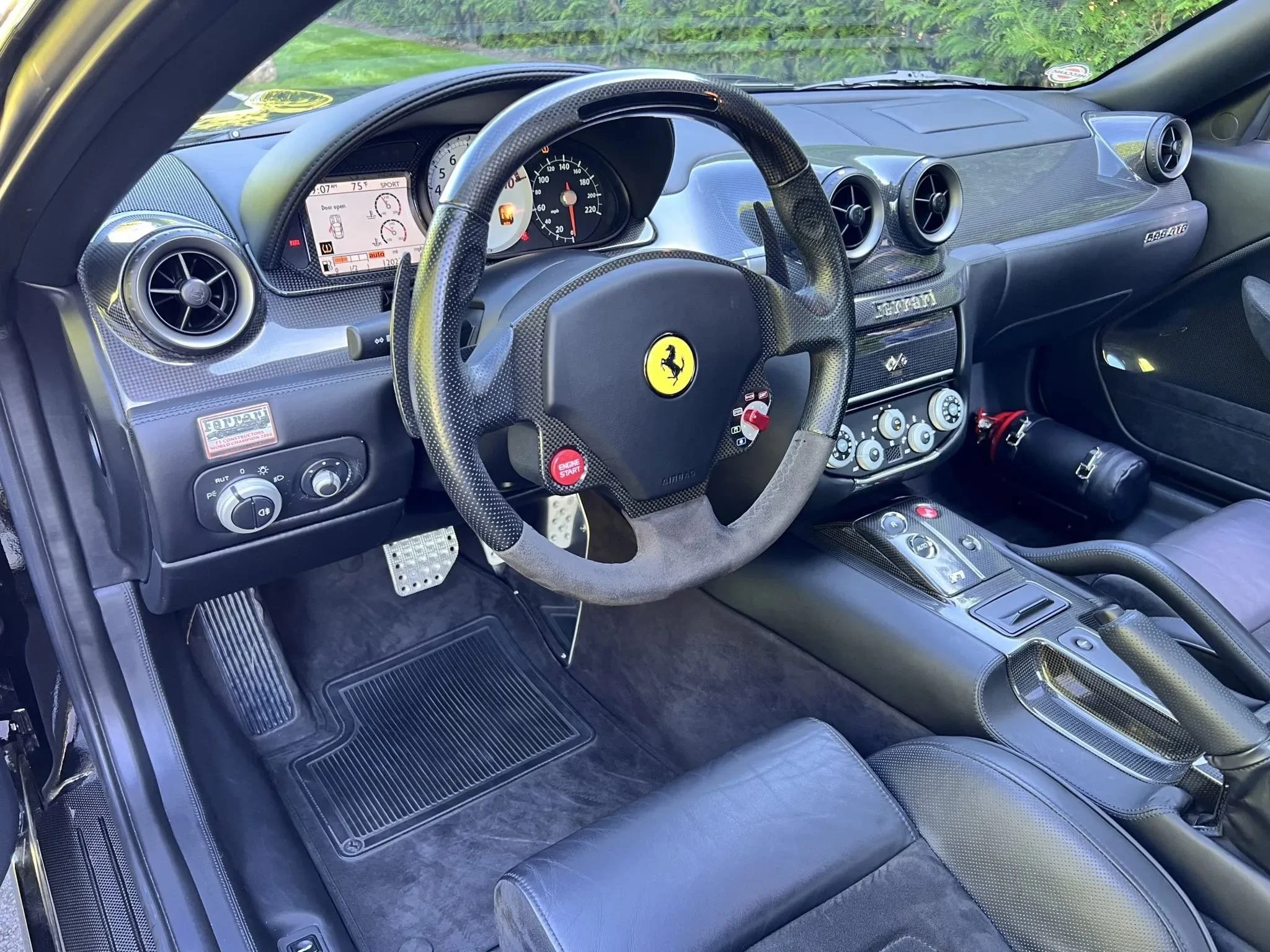 Interior of a Ferrari sports car featuring a racing-style steering wheel with a yellow Ferrari logo, digital dashboard, carbon fiber accents, and a sleek console with controls.