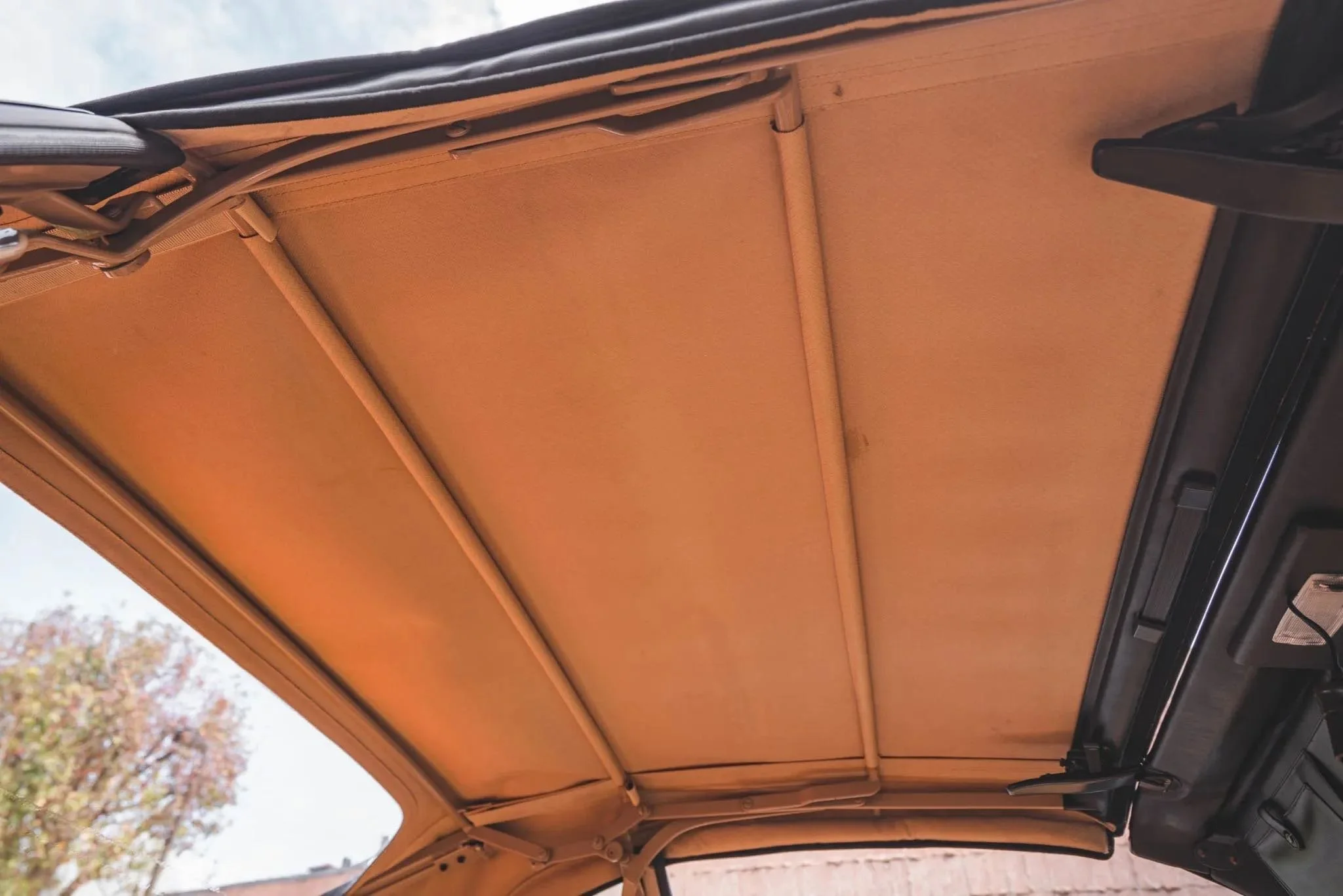 The interior roof of a vehicle with a tan or beige fabric headliner, steel support rods, and a black dashboard visible at the bottom.