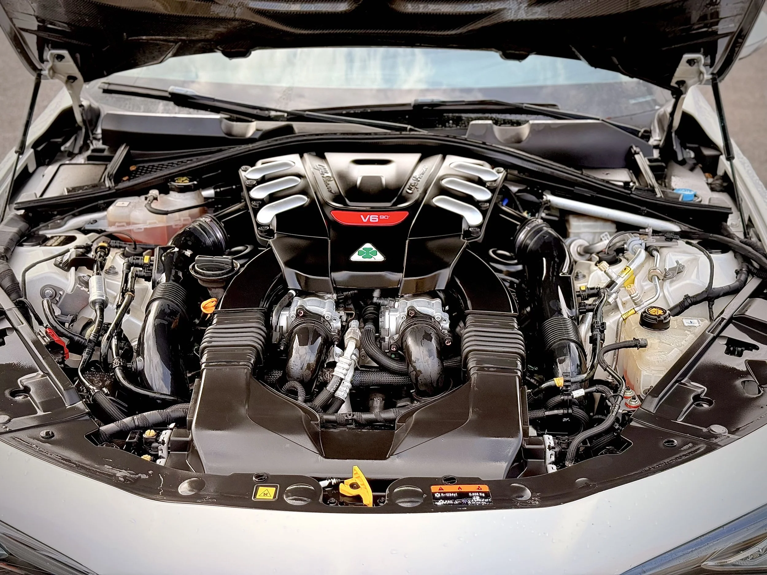 The image shows the engine bay of a sports car, featuring a V6 engine with a red label, surrounded by various mechanical components, hoses, and fluid reservoirs.