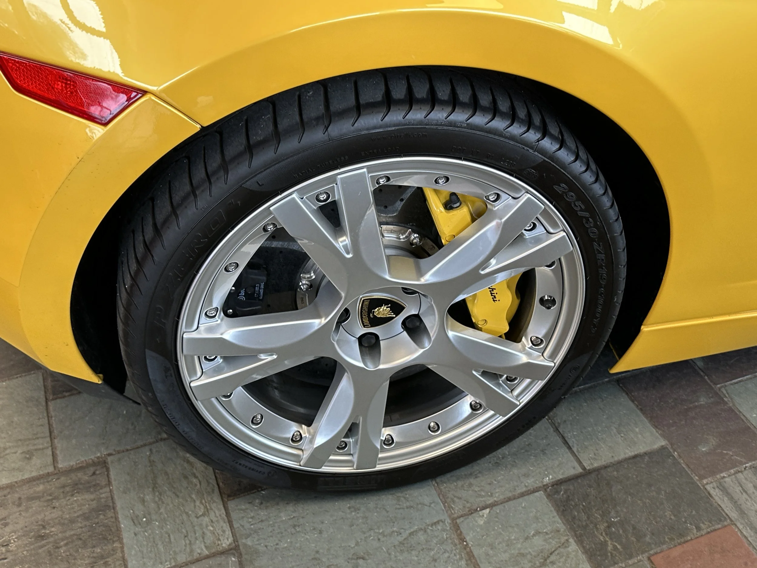 Close-up of a yellow sports car wheel showing a silver rim, yellow brake caliper, tire, and a yellow car body with a brake light.
