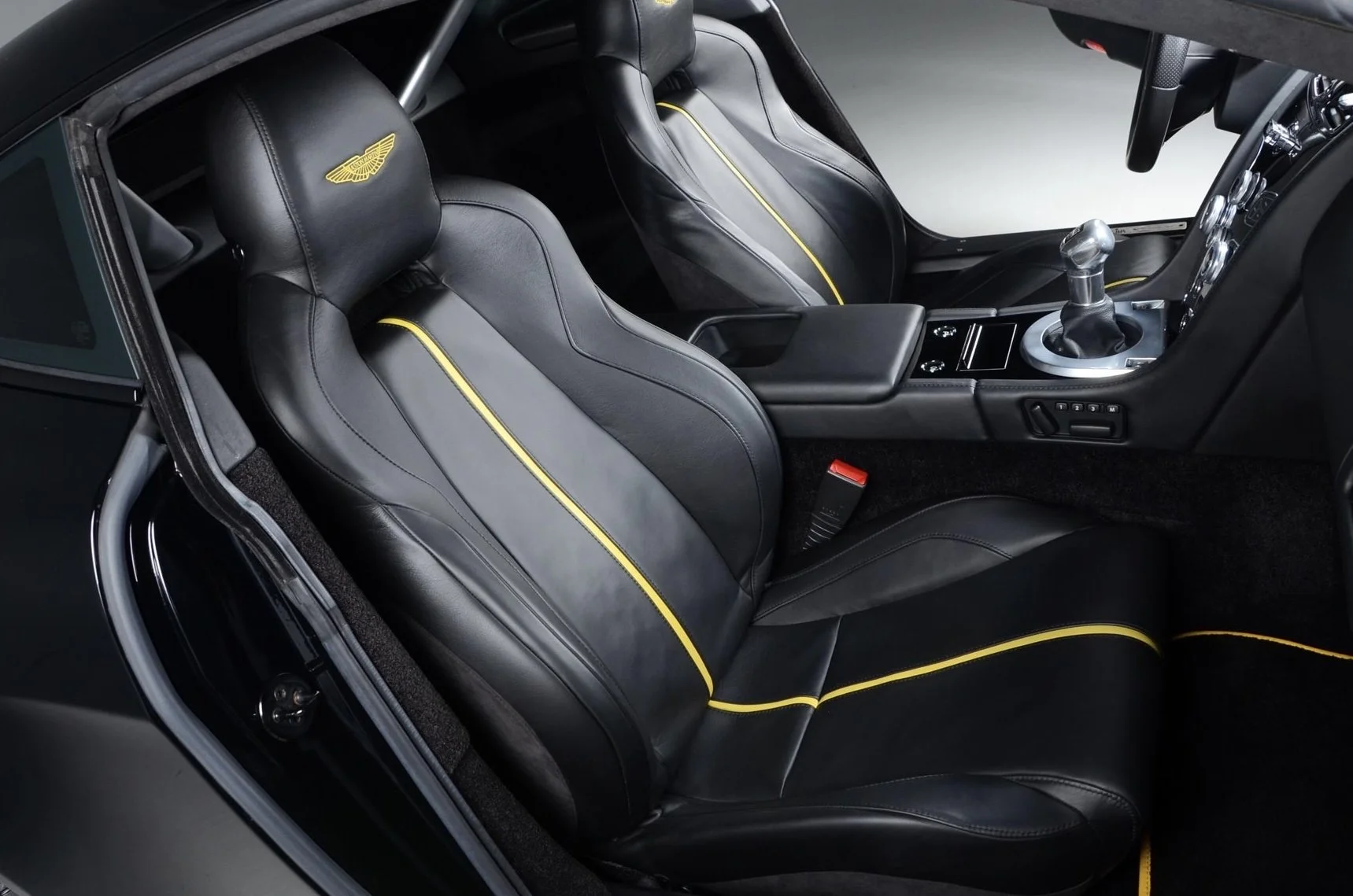 The interior of a luxury sports car featuring black leather seats with yellow piping and an Aston Martin logo on the headrests. The image captures the driver's and passenger's seats, center console with gear shifter, and part of the dashboard.