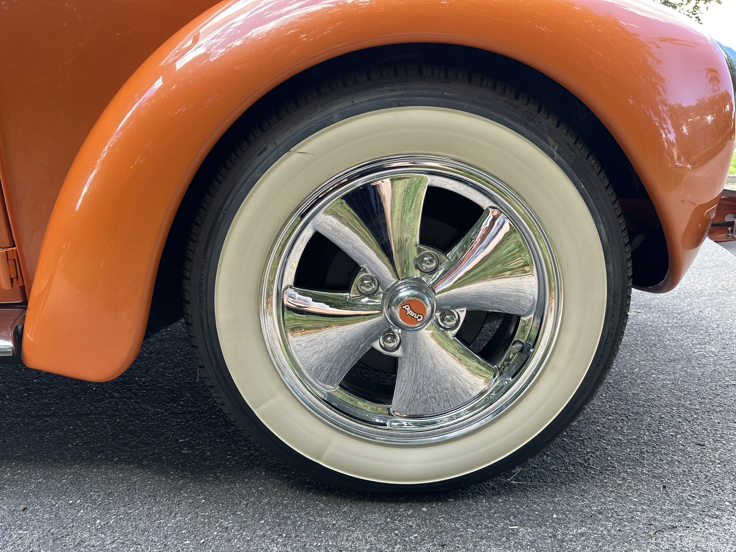 Close-up of a vintage car wheel featuring a shiny chrome hubcap with a red center logo, cream whitewall tire, orange fender, and a reflections of greenery and sky.