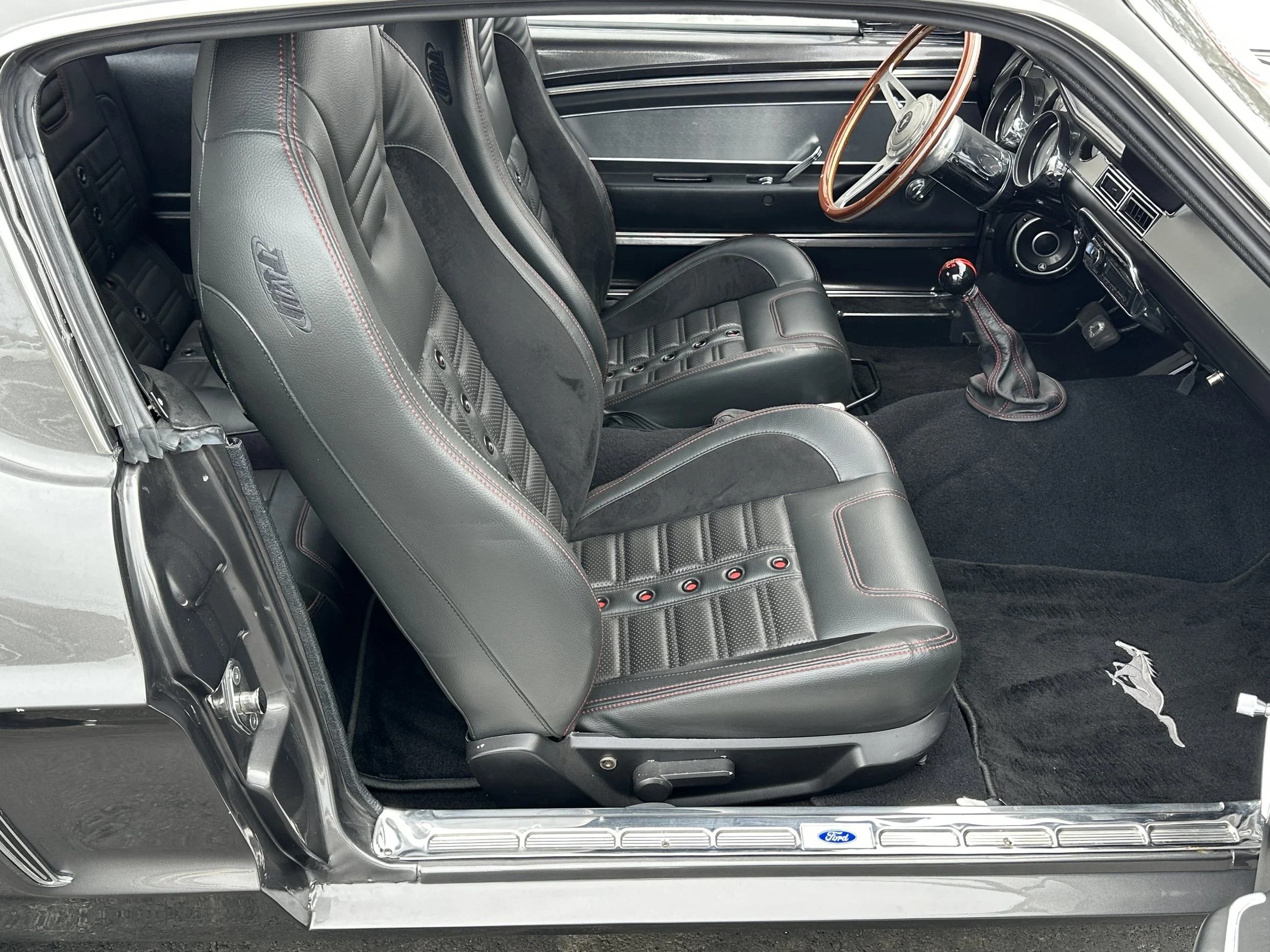 Interior of a vintage Ford Mustang, featuring black leather seats with red stitching, a wooden steering wheel, a gear shifter with a red top, and a black carpet with a Mustang logo.