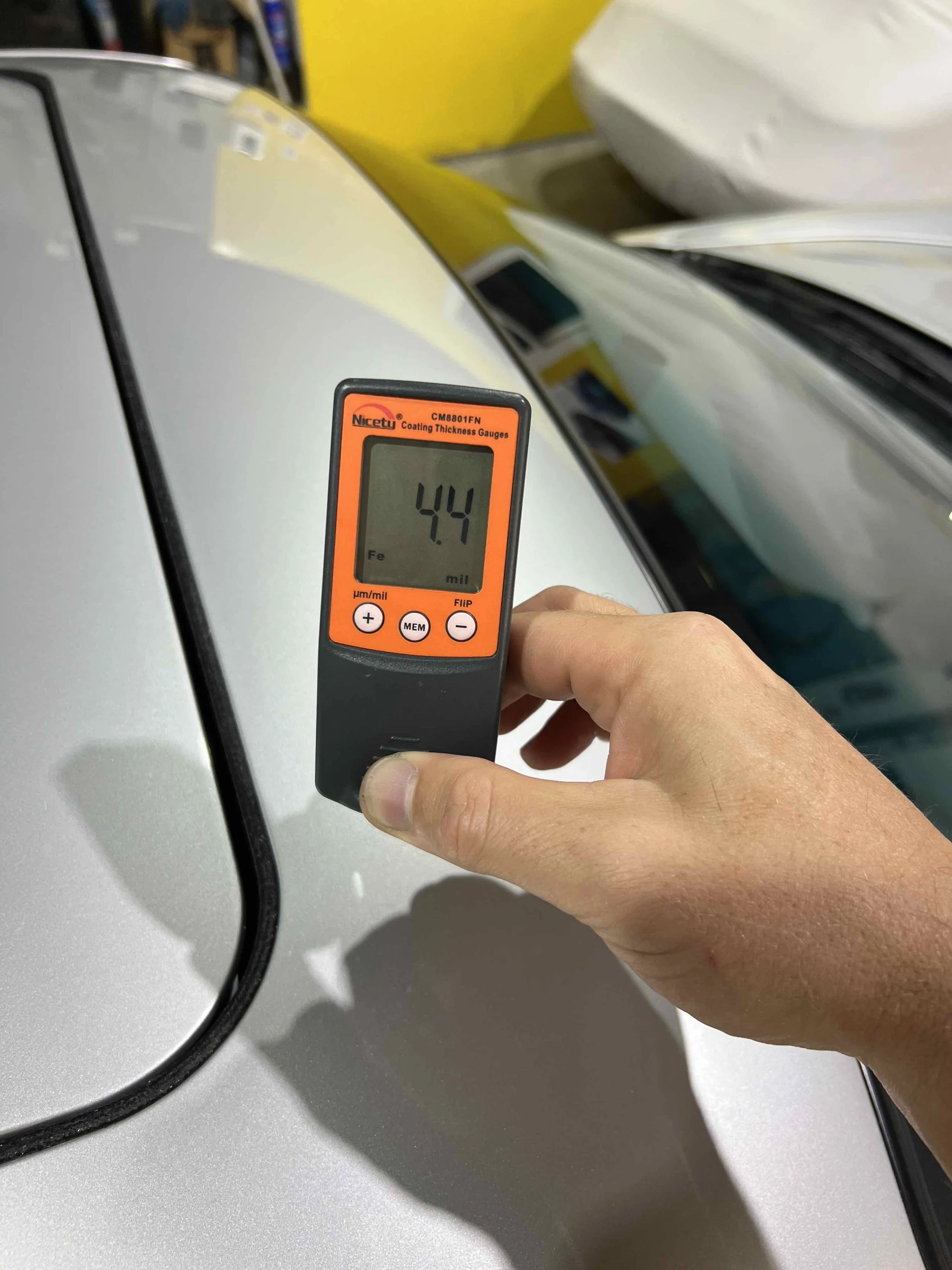 A person holding a coating thickness gauge measuring the paint thickness on a silver car's surface. The gauge display shows a reading of 4.4.