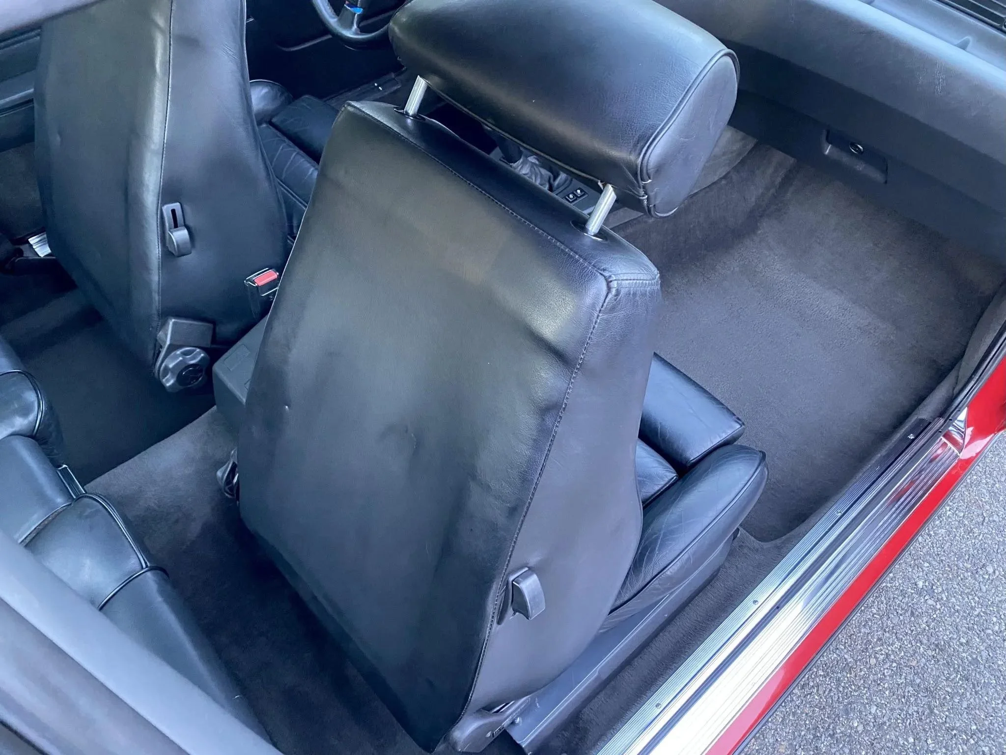 Interior of a car showing the back of the front seats, with the backside of the driver's seat and passenger seat visible, both covered in black leather, and the gray carpeted floor.
