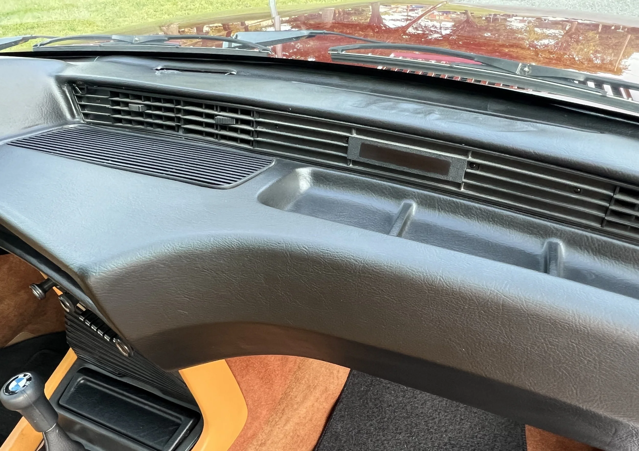 Close-up of a vintage BMW 630 csi car dashboard with black textured plastic, a black air vent, and a small compartment, with part of a yellow gear shift and brown carpet visible.