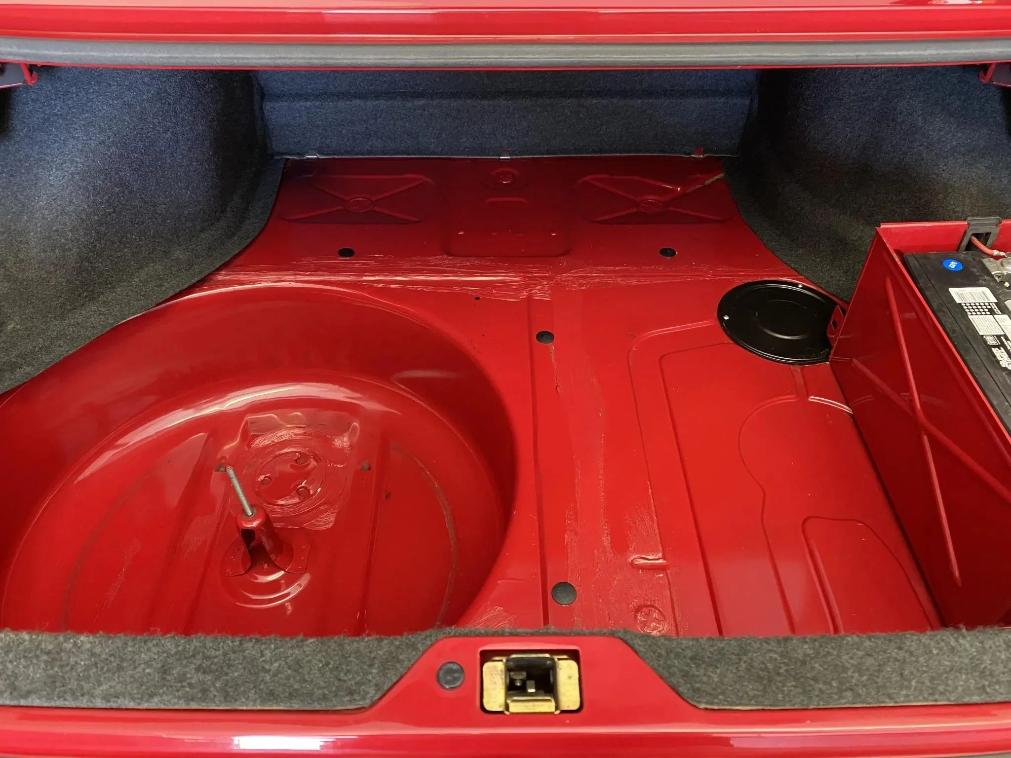 The trunk of a red car with a spare tire well and space for a tire, along with a small battery compartment on the right side.