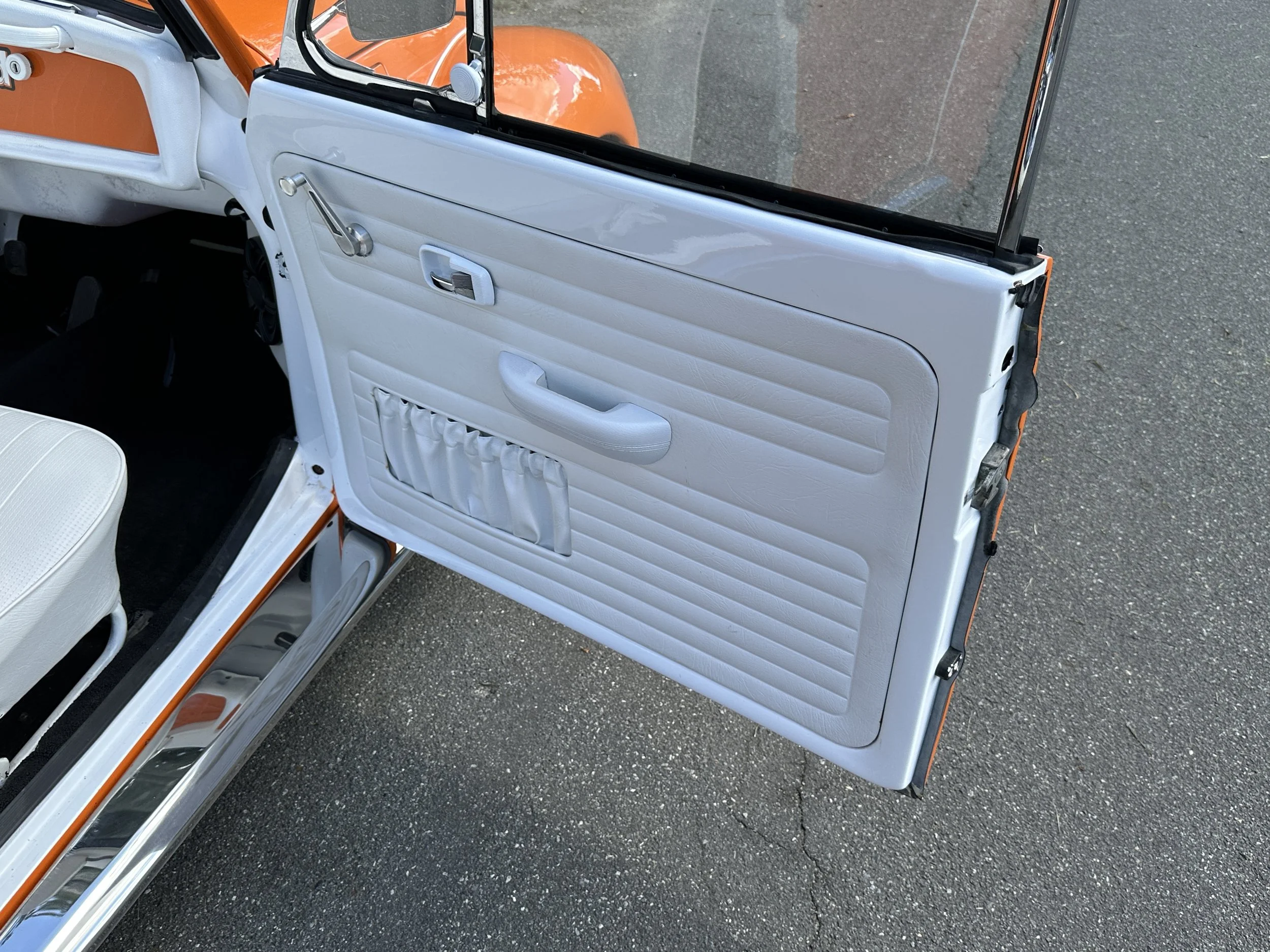 Interior of a vintage car featuring the driver’s side door with a white panel, window crank, door handle, and fabric pocket, parked on a paved surface.