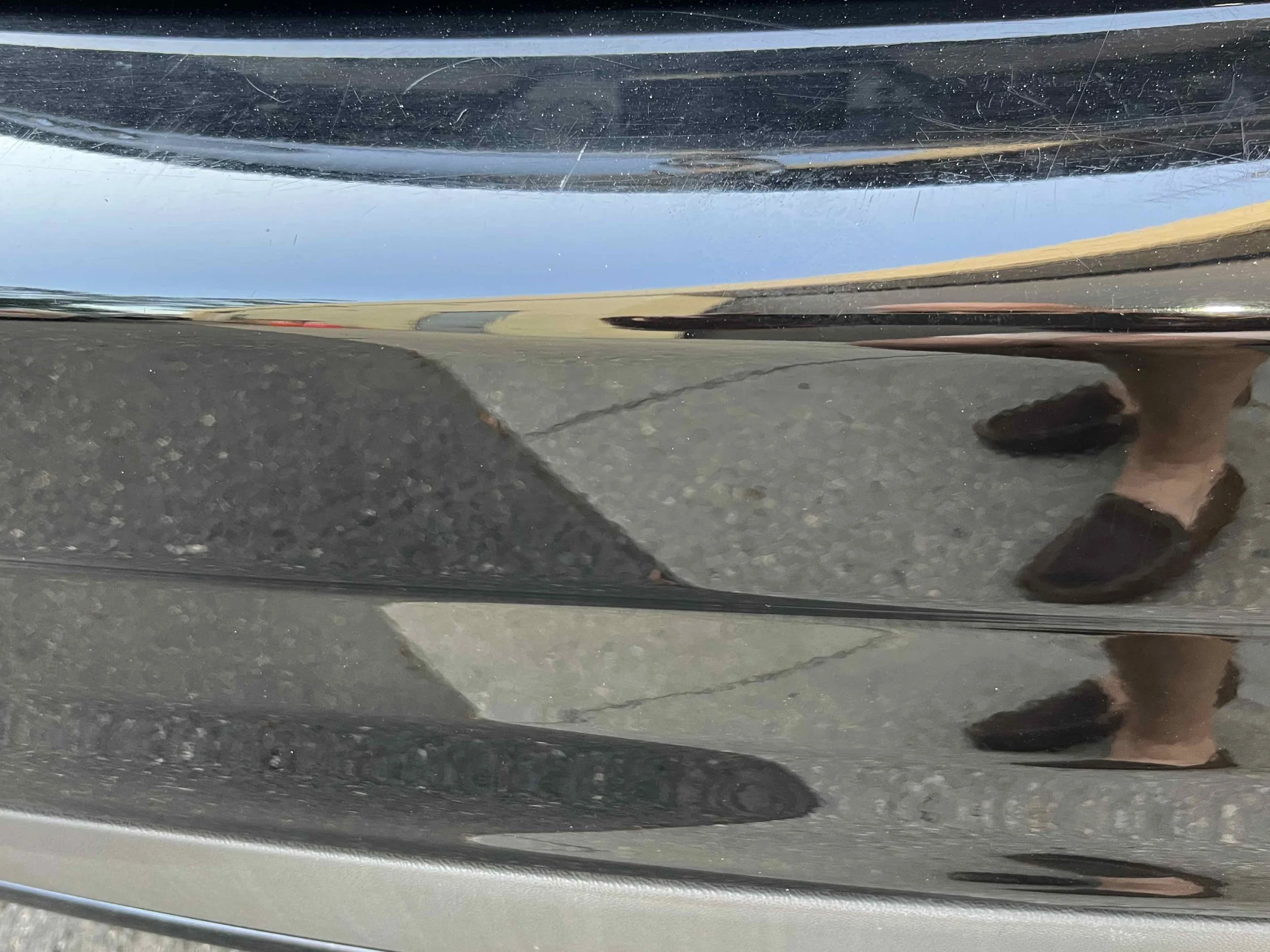 Close-up of a black car's bumper without visible scratches and scuff marks. The reflection of a person wearing black shoes and shorts is partially visible on the bumper.