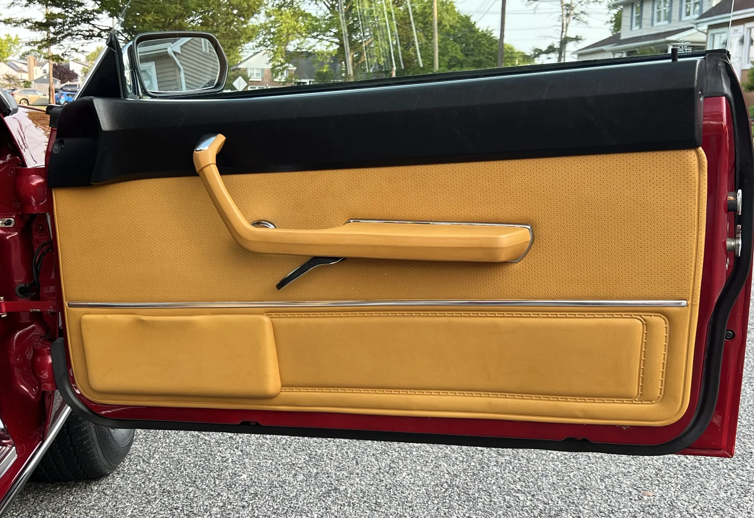 Close-up of a vintage 1977 BMW 630 csi car door interior with tan upholstery, black upper trim, and a yellow door handle, with houses and trees visible in the background.