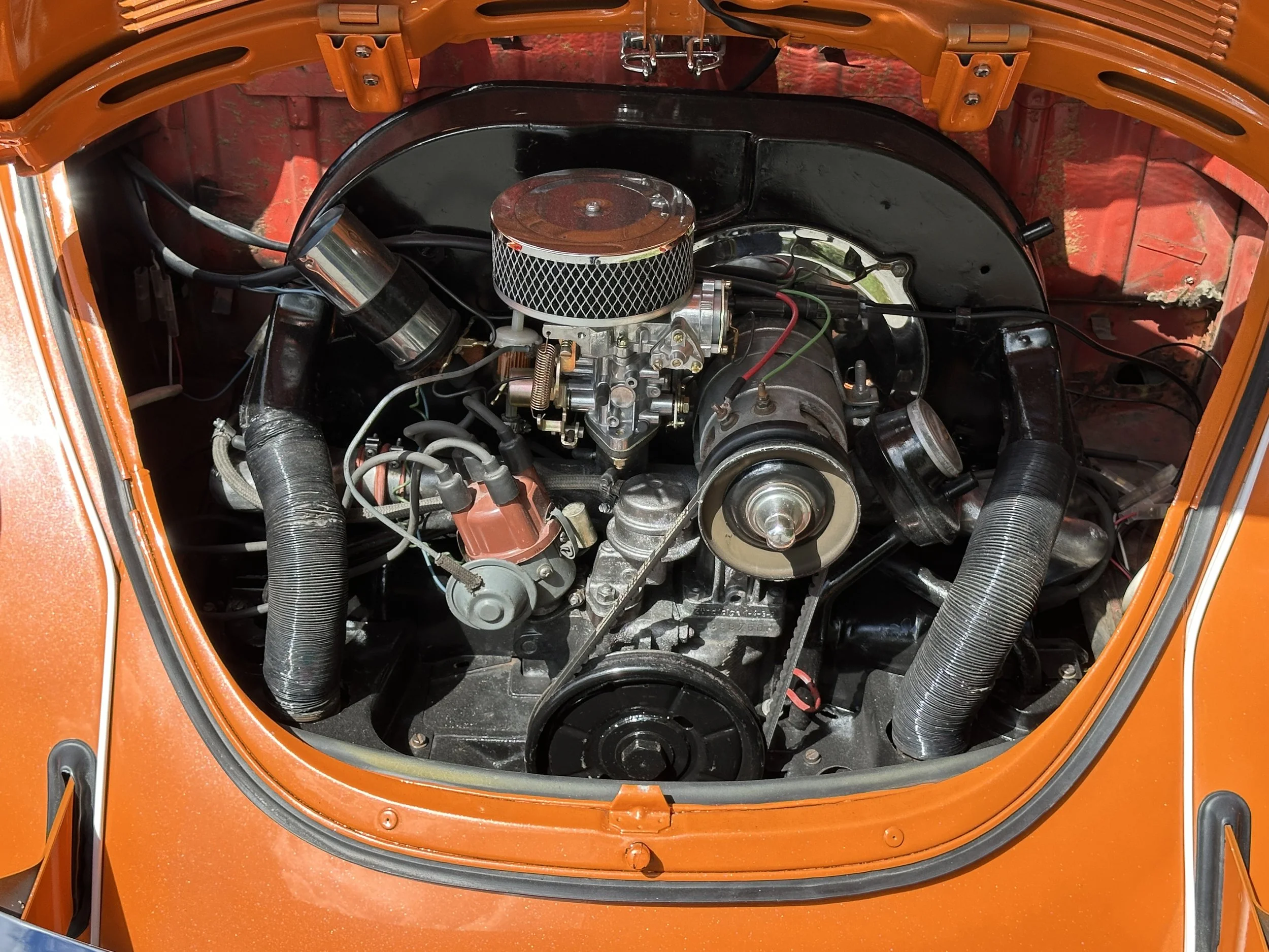 The image shows the engine bay of an orange vintage car with a V8 engine, metallic components, a black air filter, and various hoses and wires.
