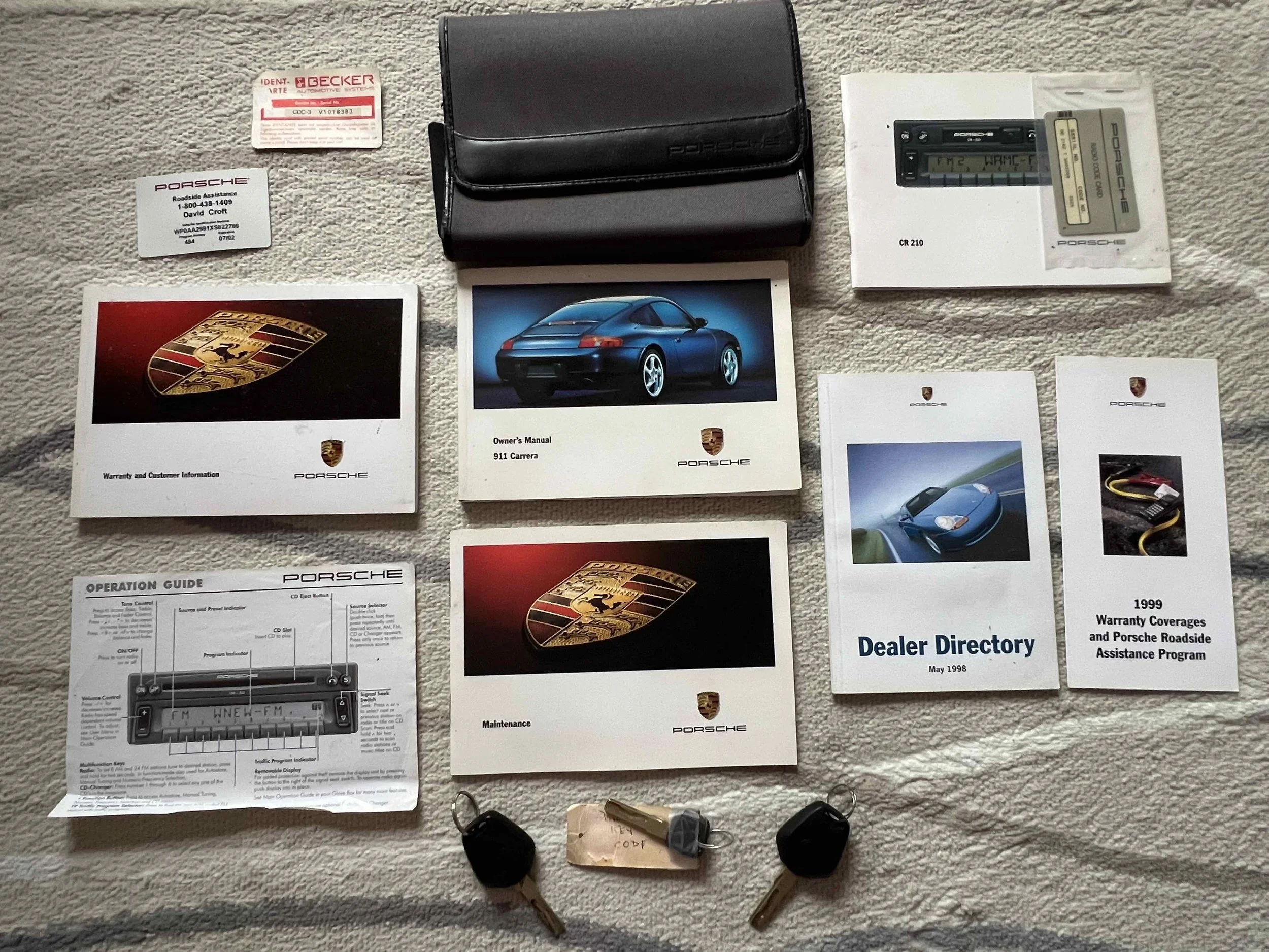 Collection of Porsche car manuals, warranty cards, a dealer directory, brand brochures, a car key, remote, and a car accessory kit on a carpeted surface.