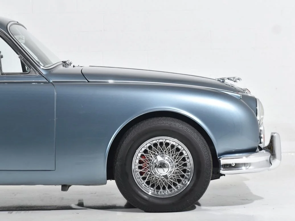 Close-up of the front half of a classic blue vintage car with wire-spoke wheels, chrome bumper, and luxury detailing against a white background.