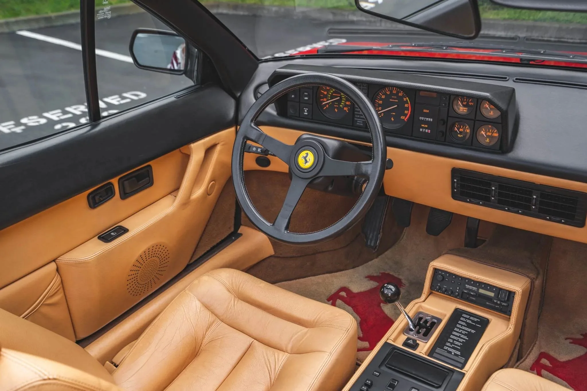 Interior of a vintage Ferrari with tan leather seats, a black steering wheel with yellow prancing horse emblem, and a dashboard with multiple gauges.